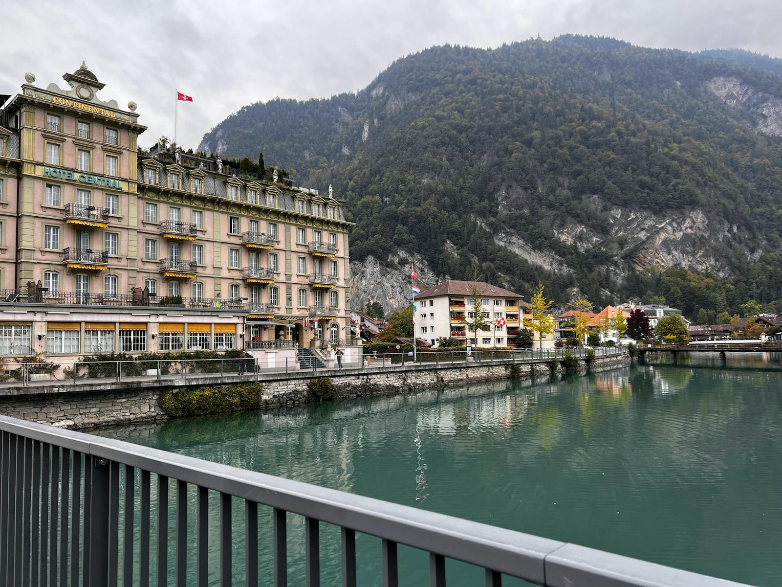 Ornate riverside hotel building beneath a forested mountain slope under overcast sky