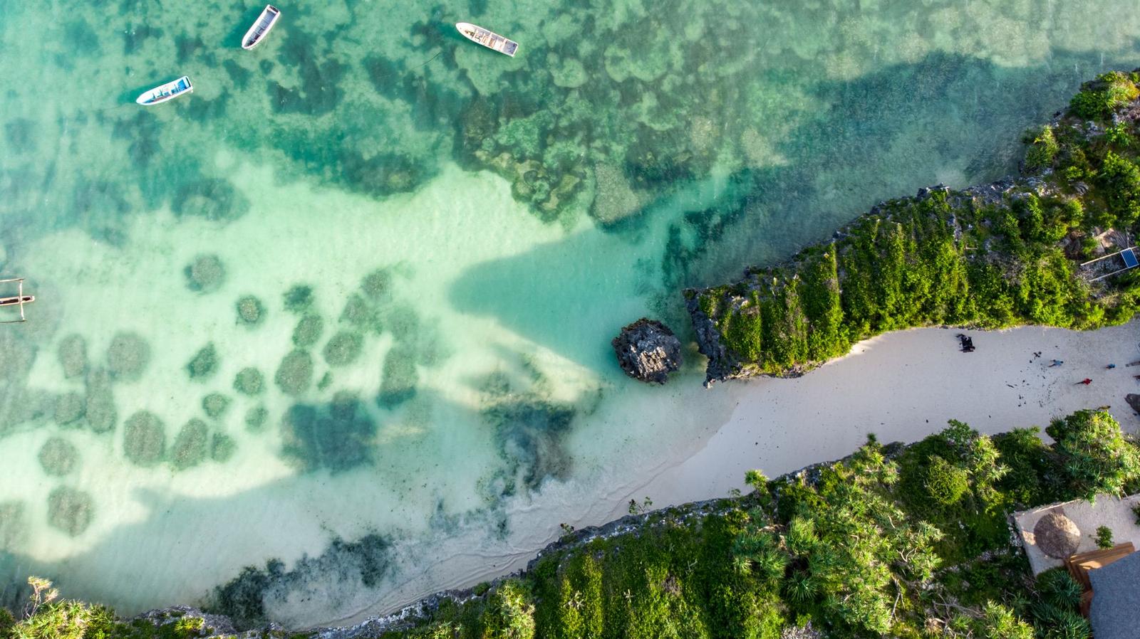 Aerial view of small boats floating over clear turquoise shallows beside a beach