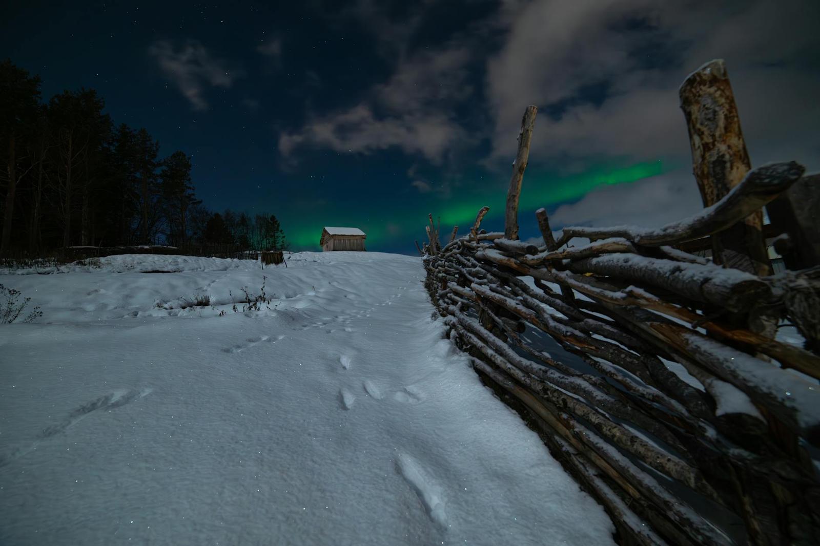 Footprints in snow beside a woven wooden fence under a faint aurora