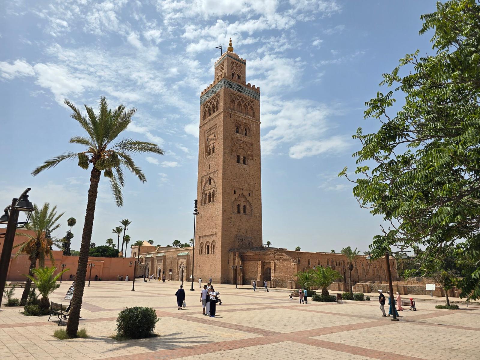 Tall sandstone minaret standing over a plaza with palm trees and strolling visitors