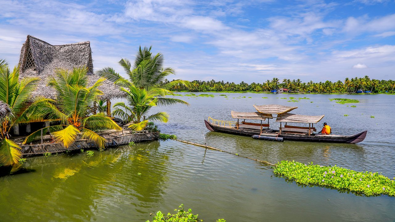 A thatched waterside hut beside a canopied boat on a palm-fringed backwater