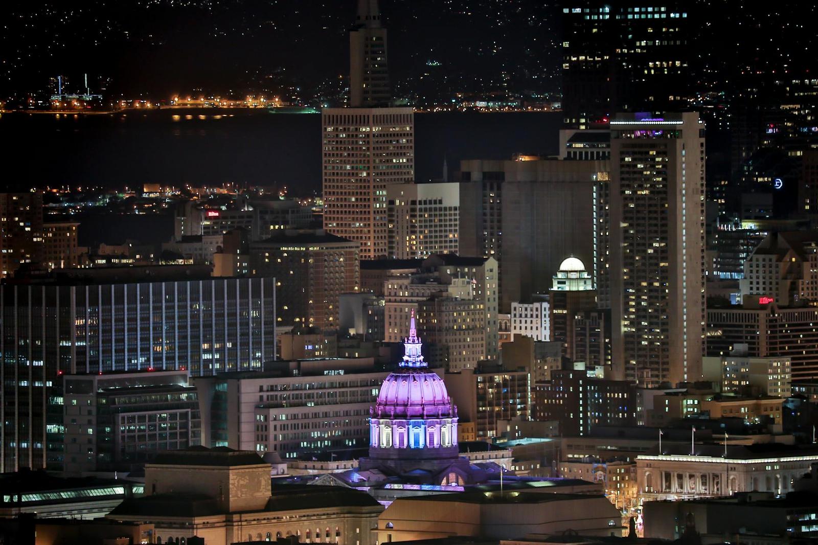 A lit city skyline at night with a domed government building glowing purple