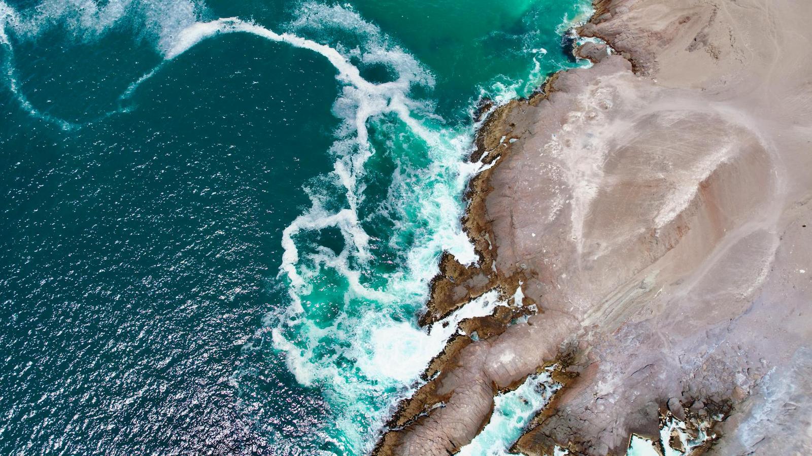 Aerial view of turquoise waves breaking against a rugged sandy coastline