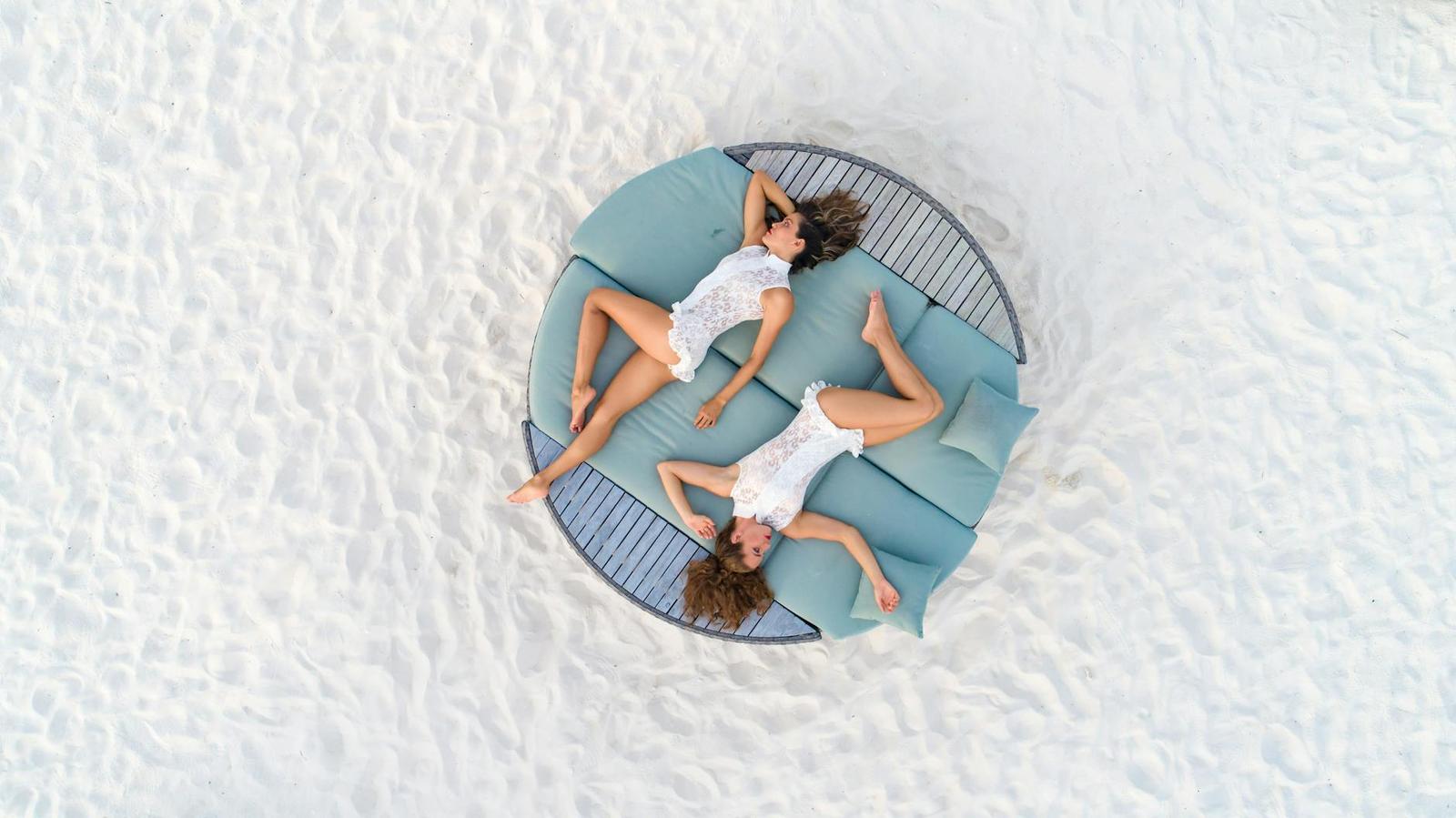 An aerial view of two women lying on a round daybed on white sand
