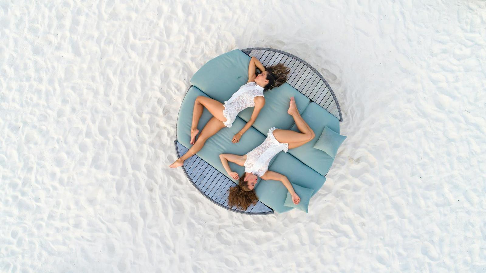An aerial view of two women lying on a round daybed on white sand
