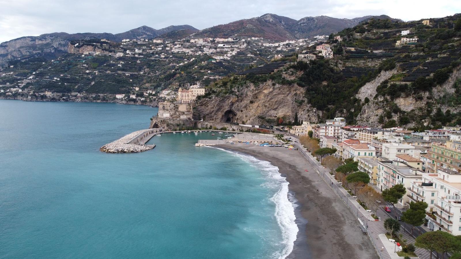 Aerial view of a curved harbour breakwater beside a pebble beach and hillside town