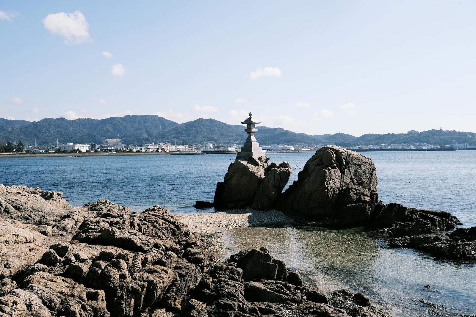 A small statue on a rocky outcrop off a coastline with hills beyond