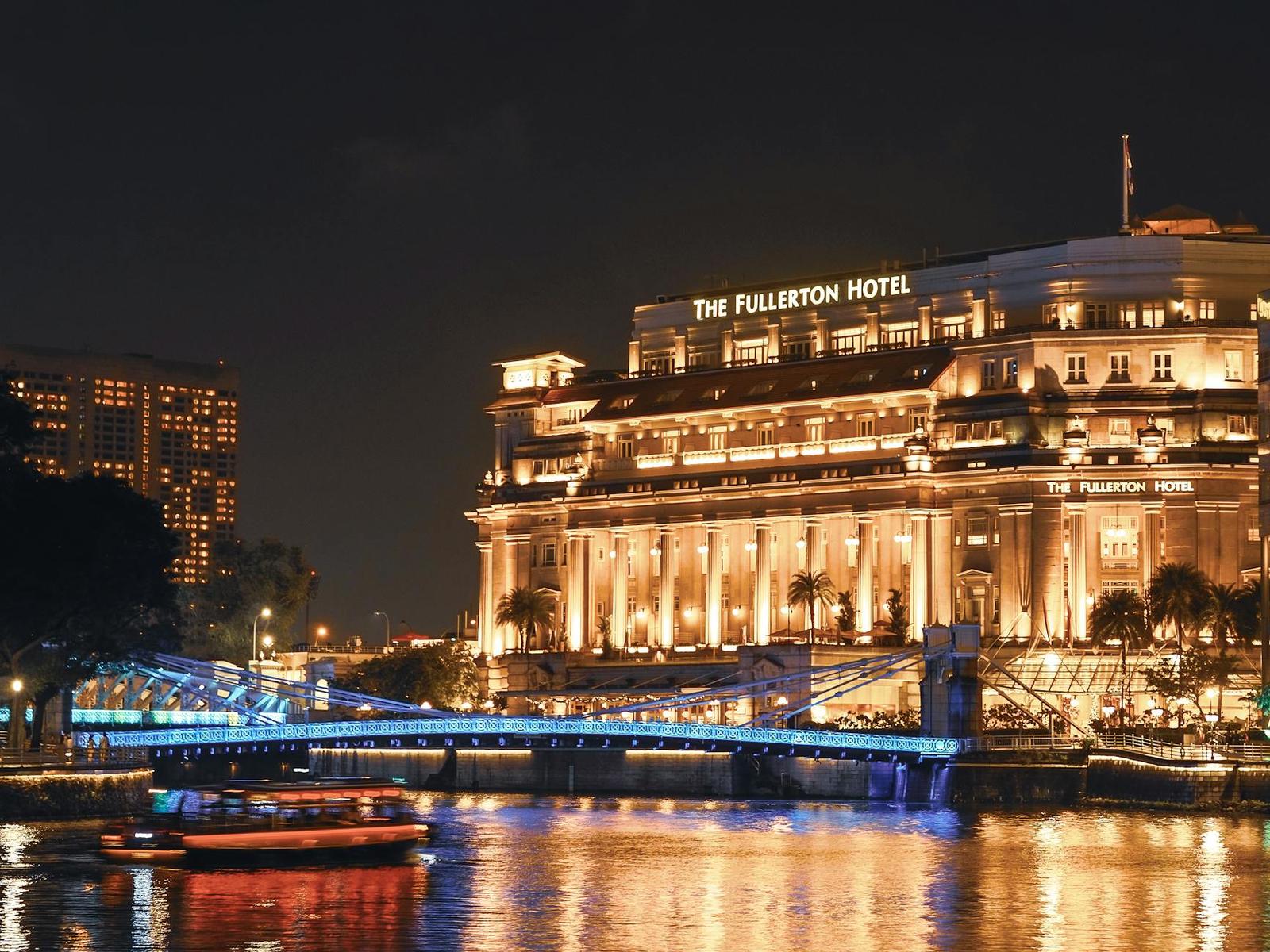 A grand illuminated colonial-style hotel beside a river with a lit footbridge at night