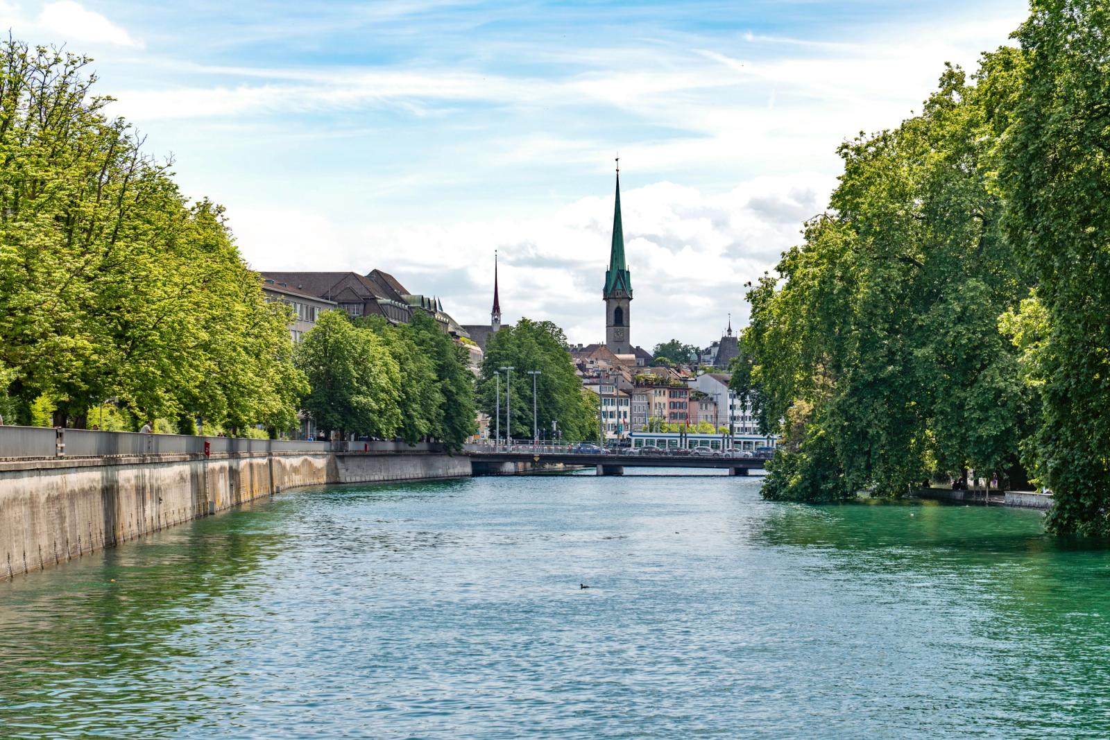 A tree-lined river running through a town toward a church spire