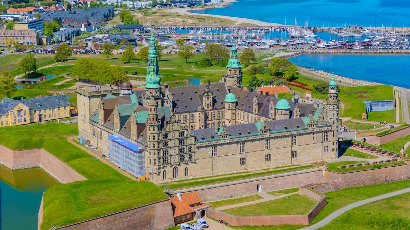 Aerial view of a moated coastal castle with green spires beside a marina
