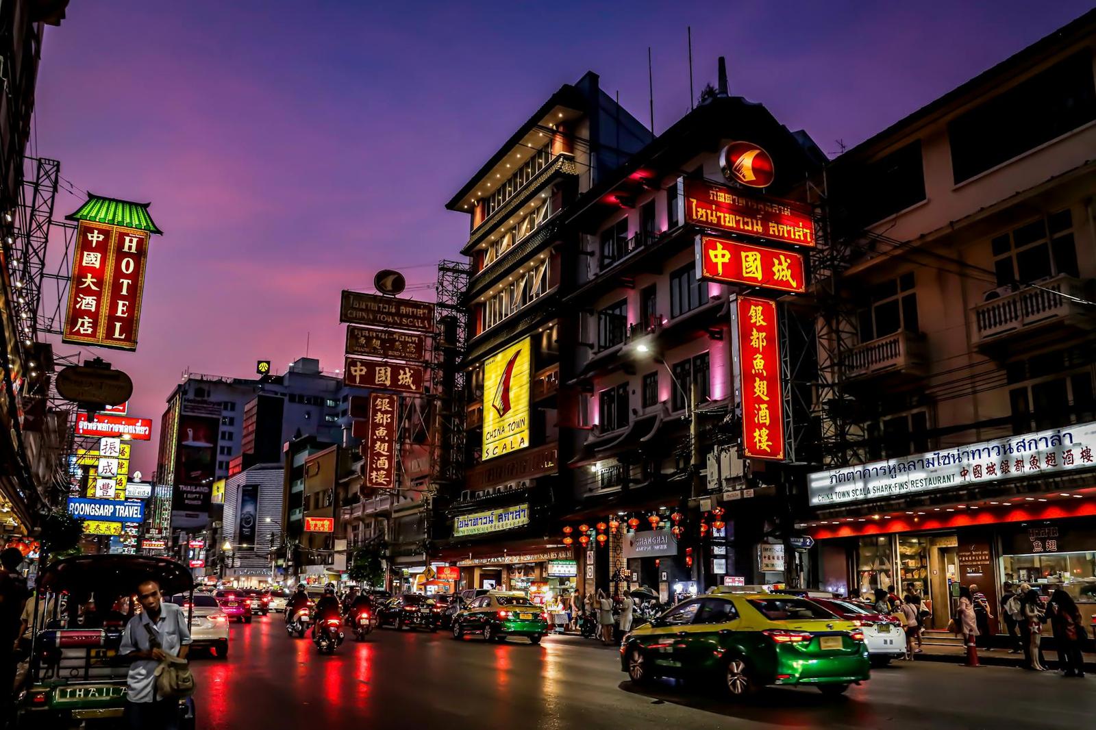 Neon-lit street at dusk with hotel signage, tuk-tuks and traffic