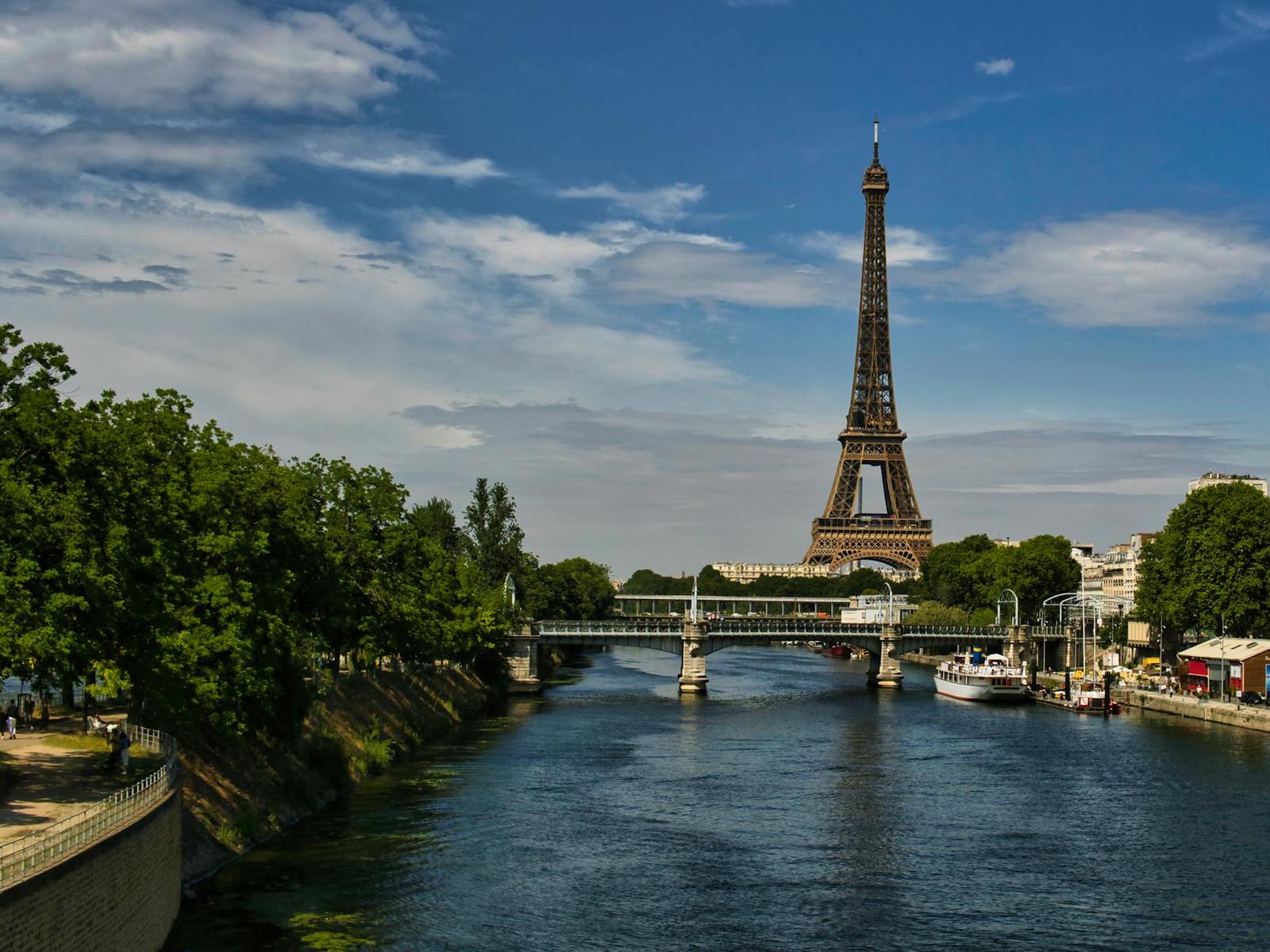 The Eiffel Tower viewed along a tree-lined river with a footbridge crossing it