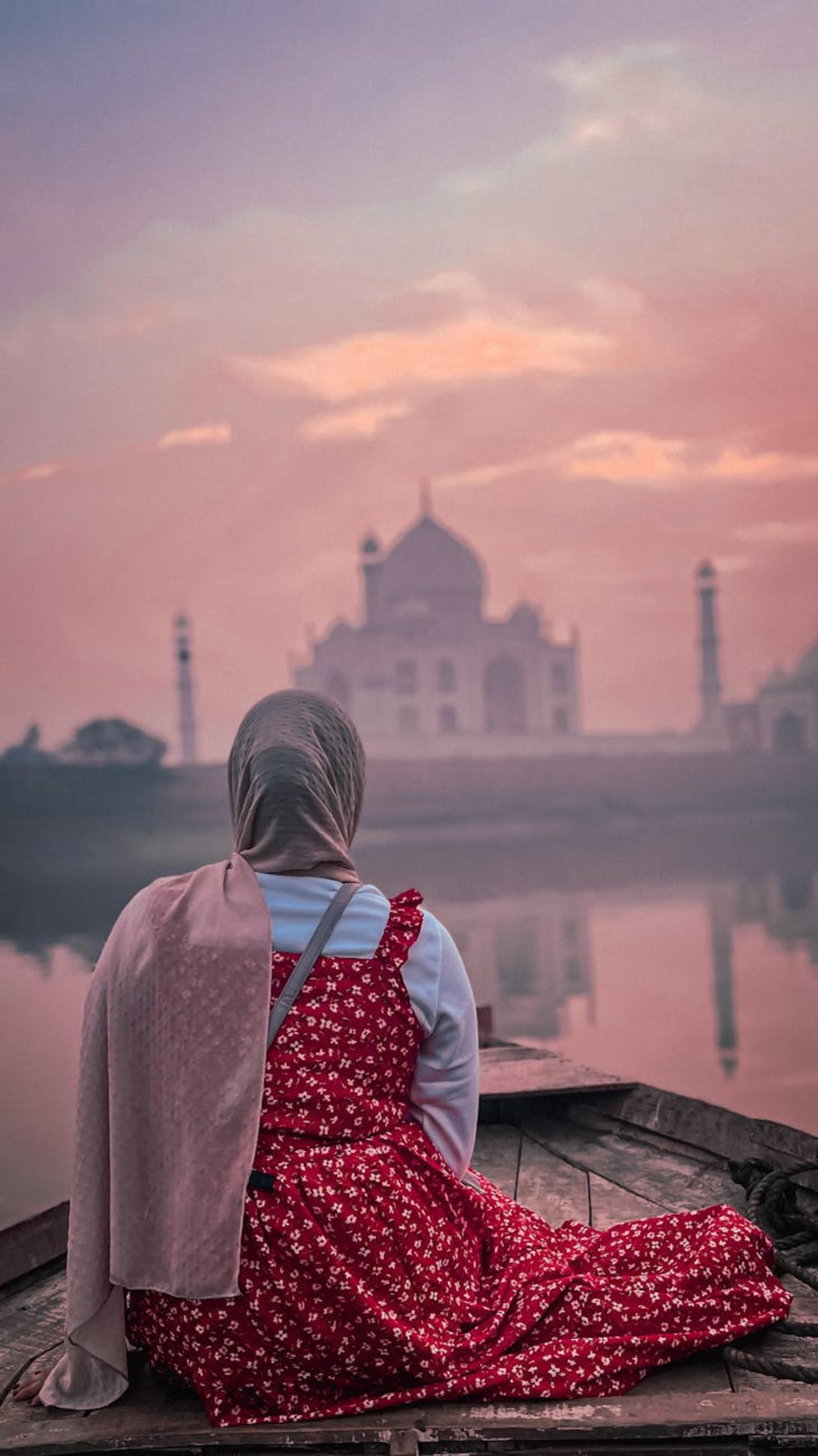 Woman in a red dress seated on a boat facing a misty domed monument at sunrise