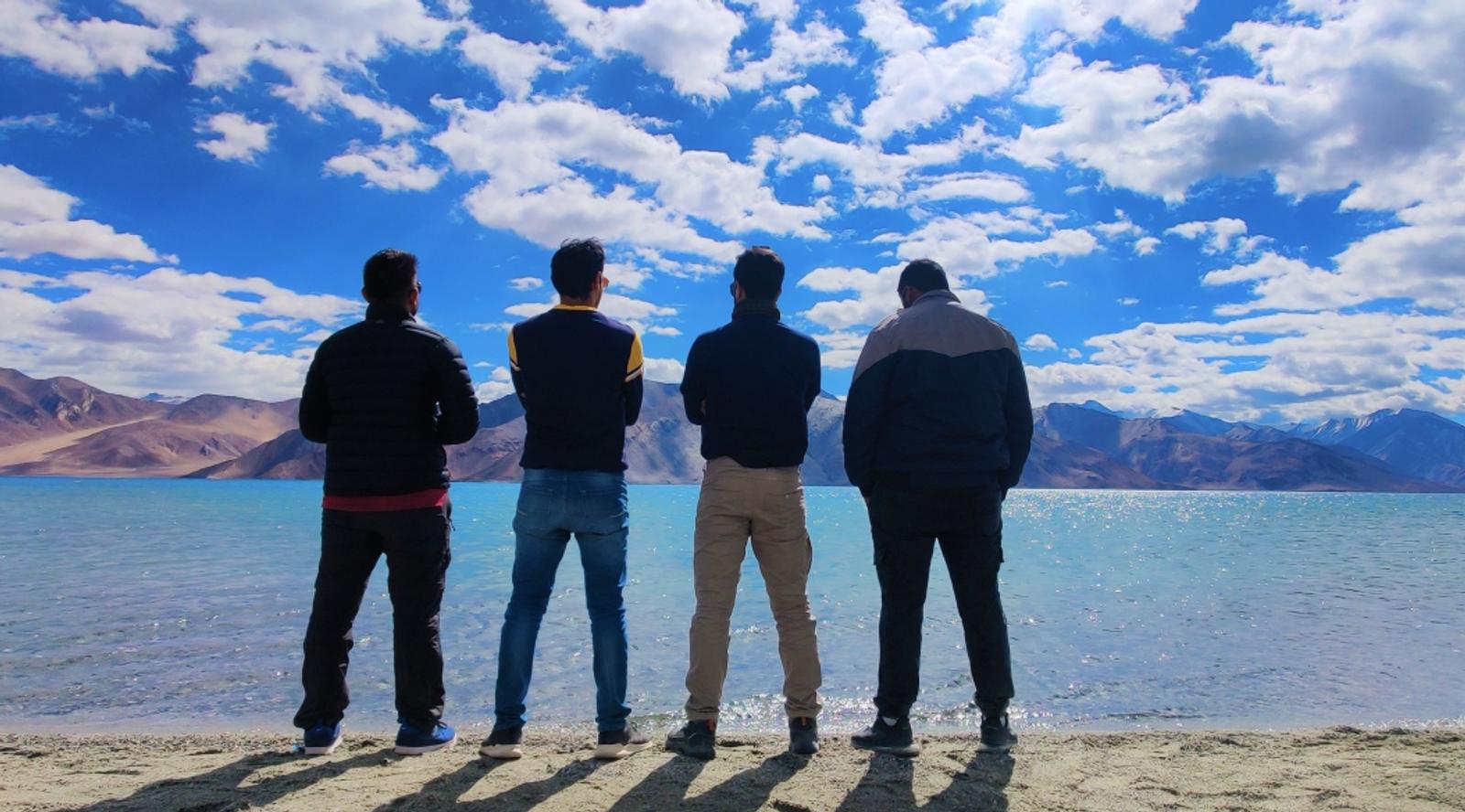 Four friends standing by a lake under a bright blue sky.