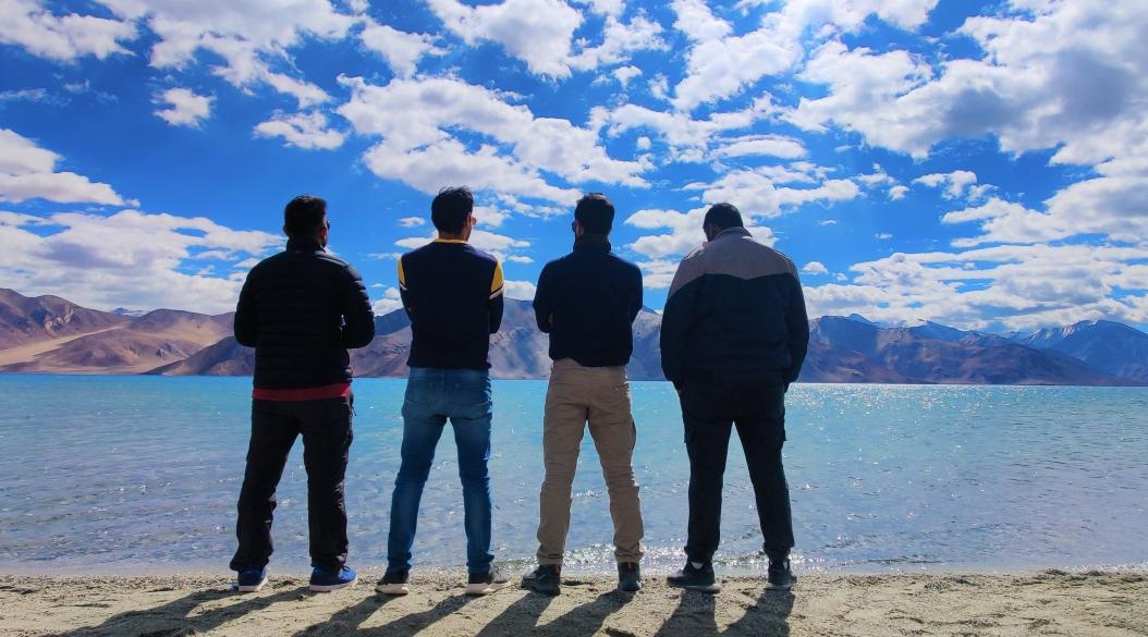 Four friends standing by a lake under a bright blue sky.