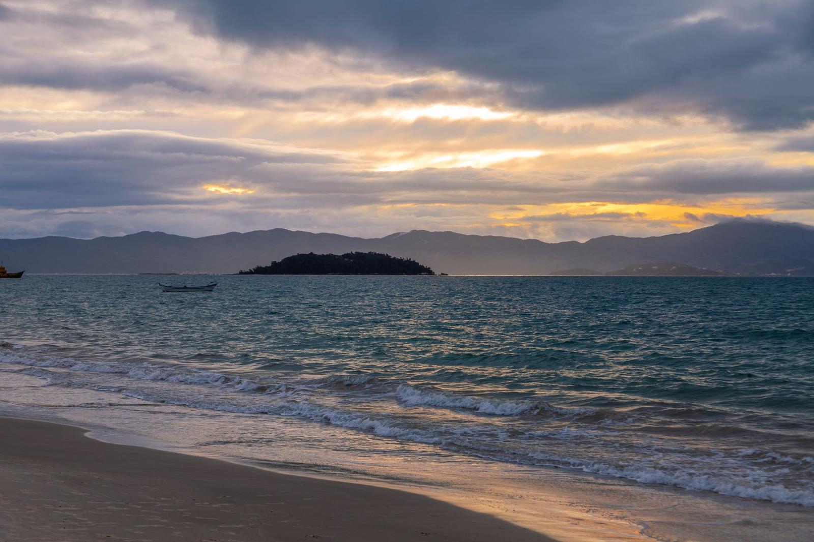 Waves wash onto a beach as a dark island silhouette rises beneath a cloudy sunset