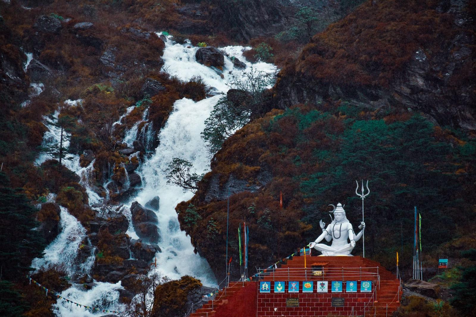 A cascading mountain waterfall beside a hilltop shrine with a statue and prayer flags