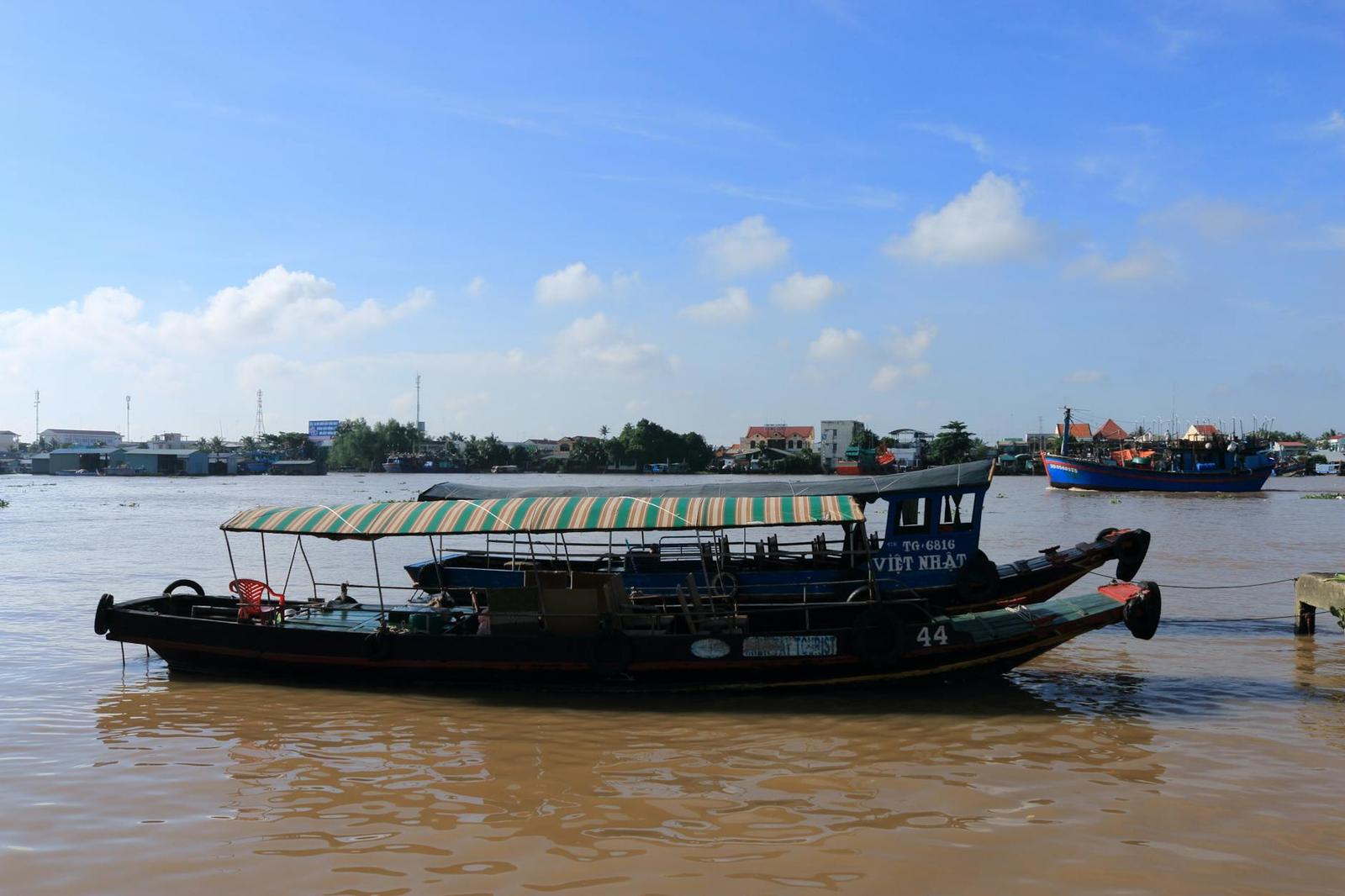A wooden riverboat with a striped canopy moored on a muddy river among other vessels