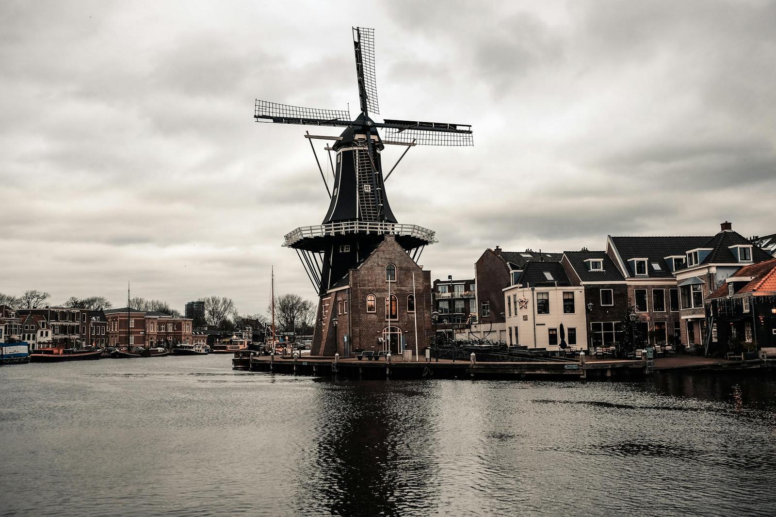 Scenic view of a traditional Dutch windmill by a river with urban buildings under cloudy skies