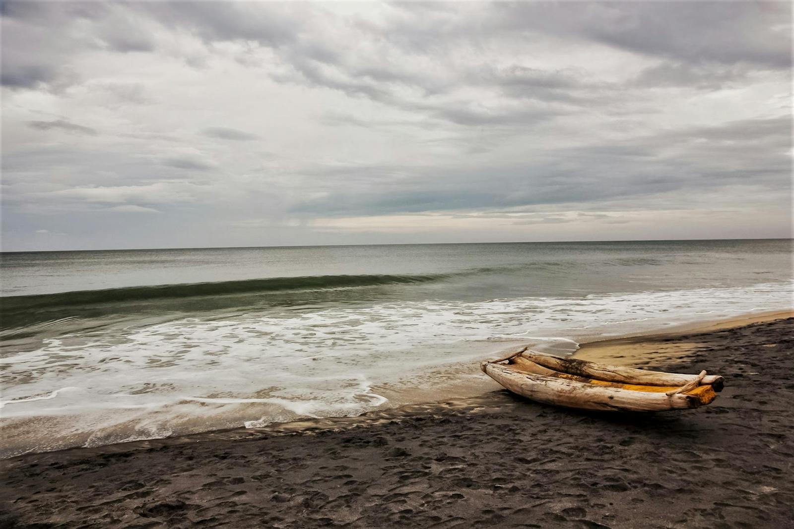 A wooden outrigger canoe resting on a dark sand beach under overcast sky