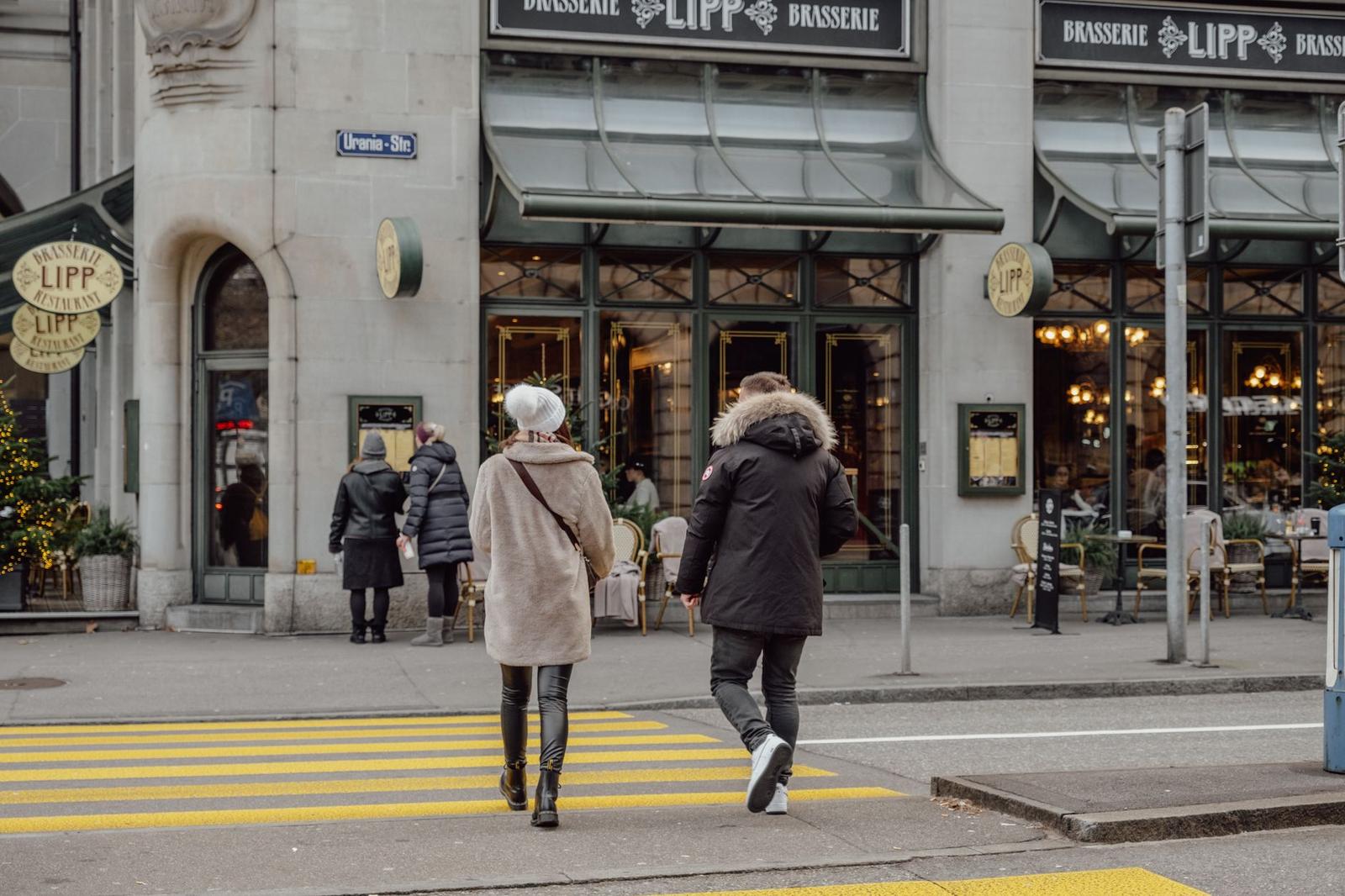 Pedestrians crossing a street lined with awning-covered brasserie cafes