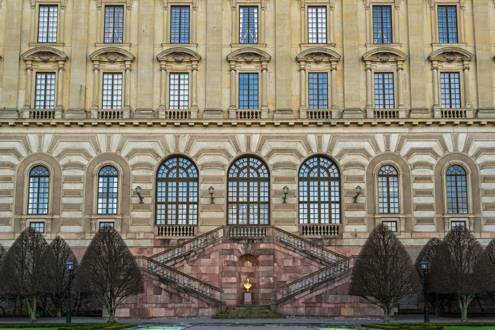 A grand baroque palace facade with a symmetrical stone staircase flanked by trimmed trees