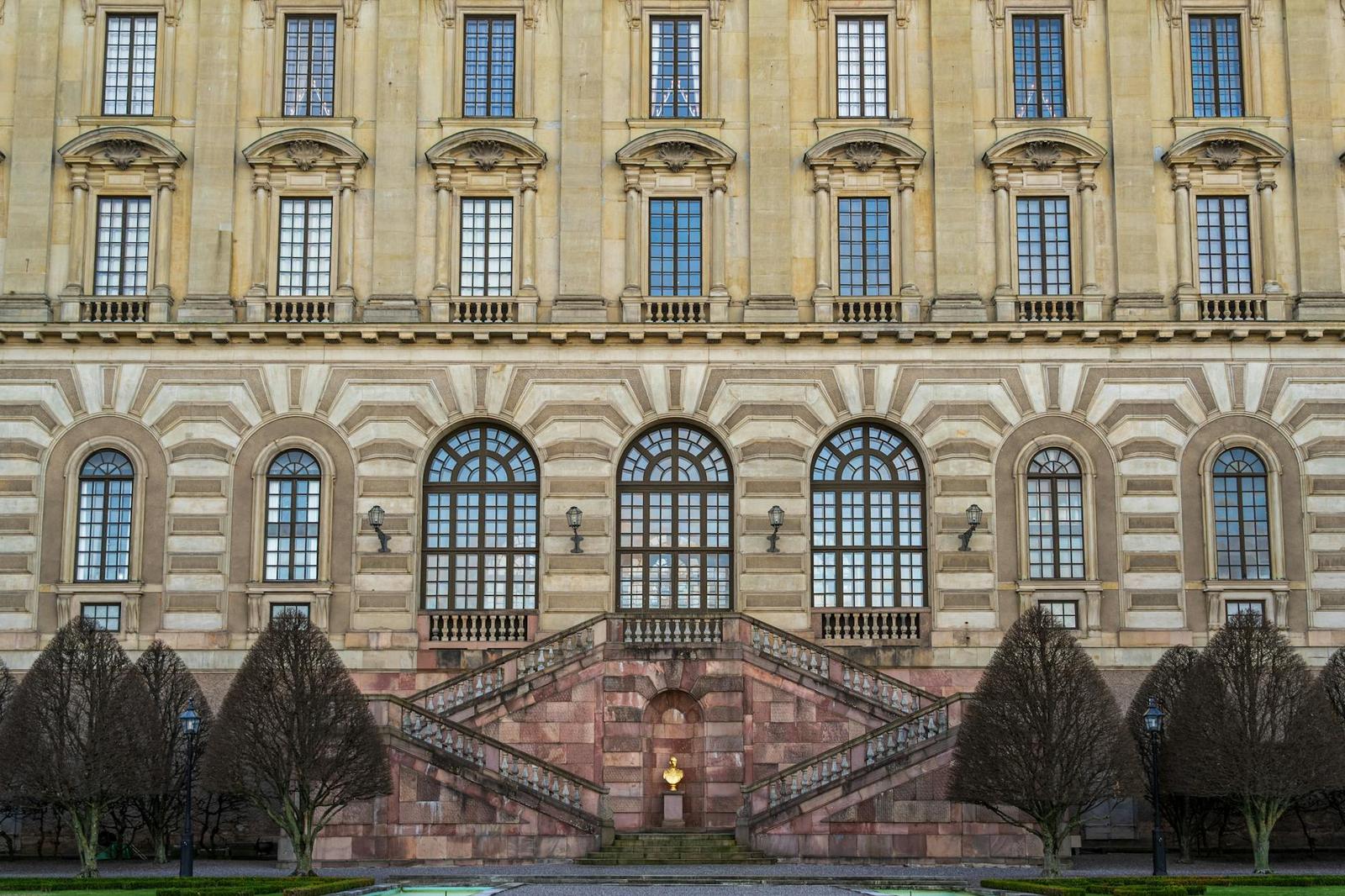 A grand baroque palace facade with a symmetrical stone staircase flanked by trimmed trees