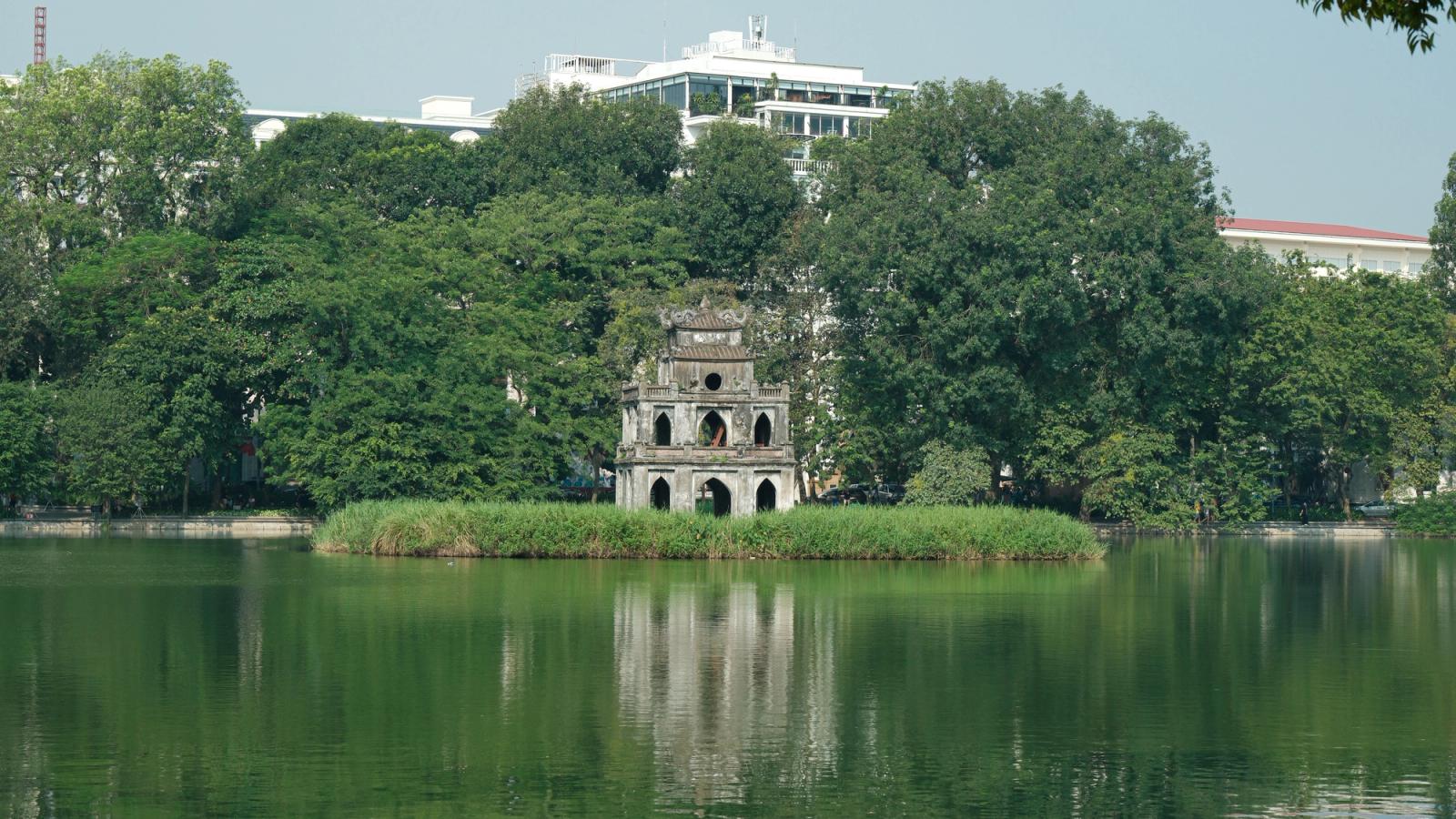Serene scene of Turtle Tower reflecting in Hoan Kiem Lake, amidst lush greenery on a sunny day