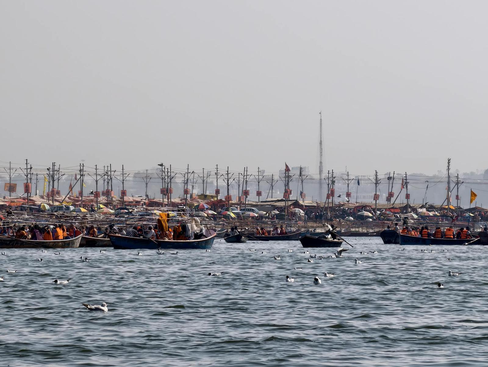 Boats crowded with pilgrims near a riverside camp of flags