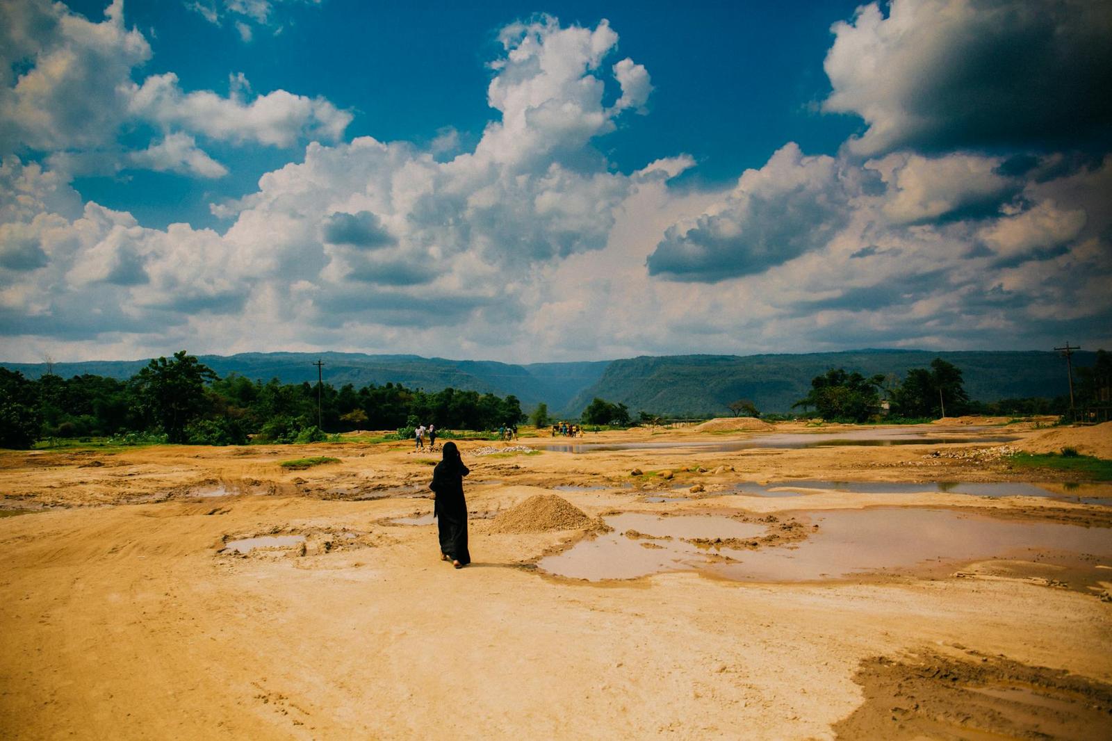 A lone figure crossing a dry ochre landscape of pools under a cloudy sky