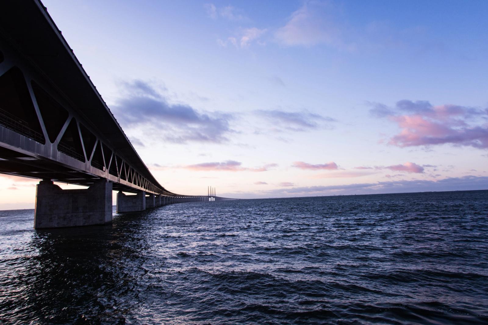 A breathtaking view of a long steel bridge stretching over a vast body of water during sunset, under a vivid sky