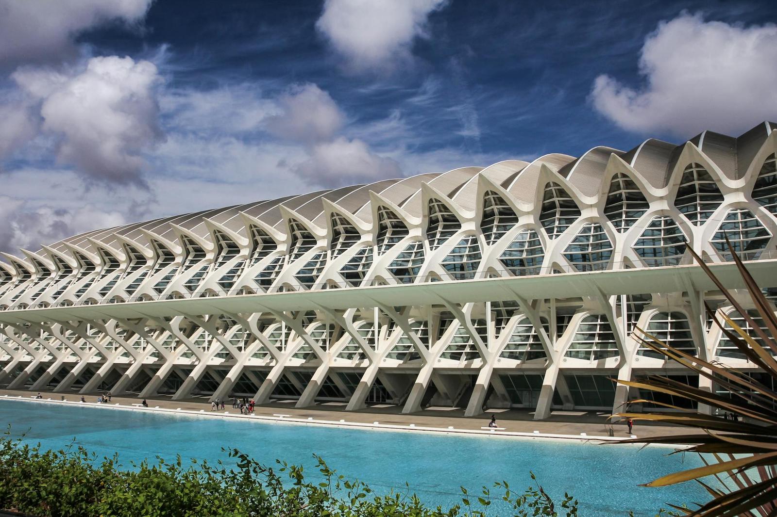 A rib-vaulted futuristic building reflected in a long turquoise pool under a cloudy sky