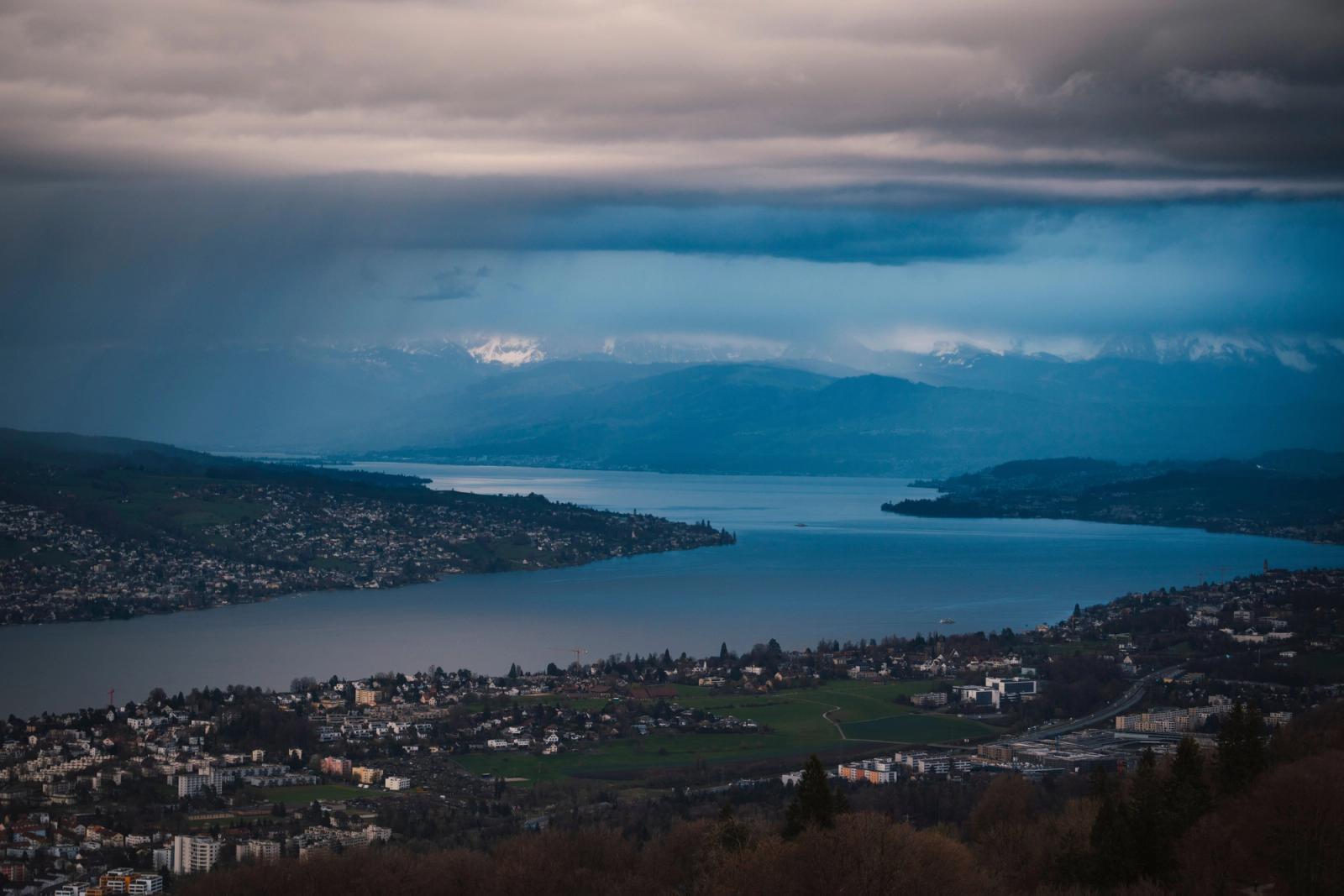 A breathtaking aerial view over Lake Zurich with distant snow-capped mountains under dramatic skies