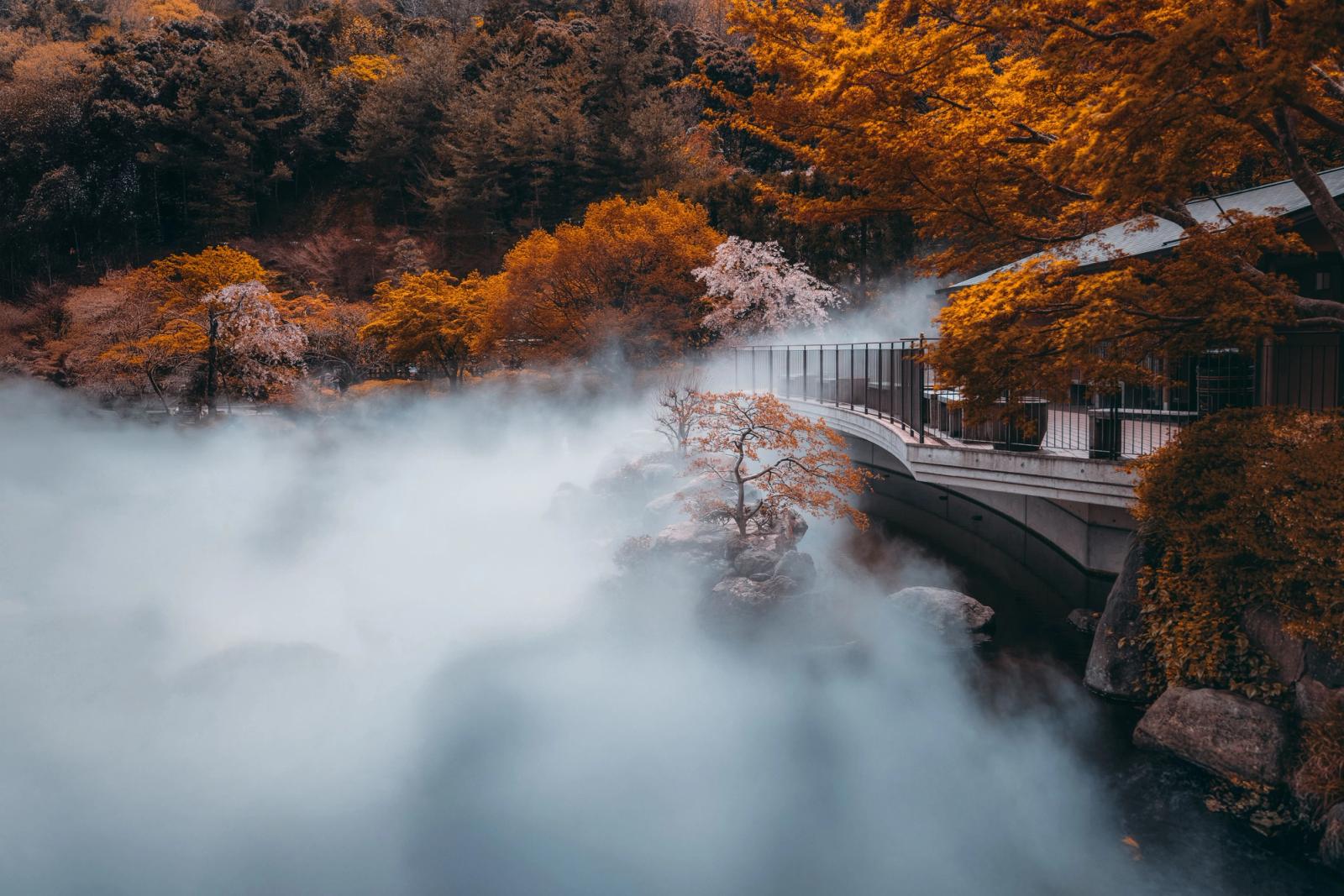 Mist rising over a river beside autumn trees and a covered footbridge