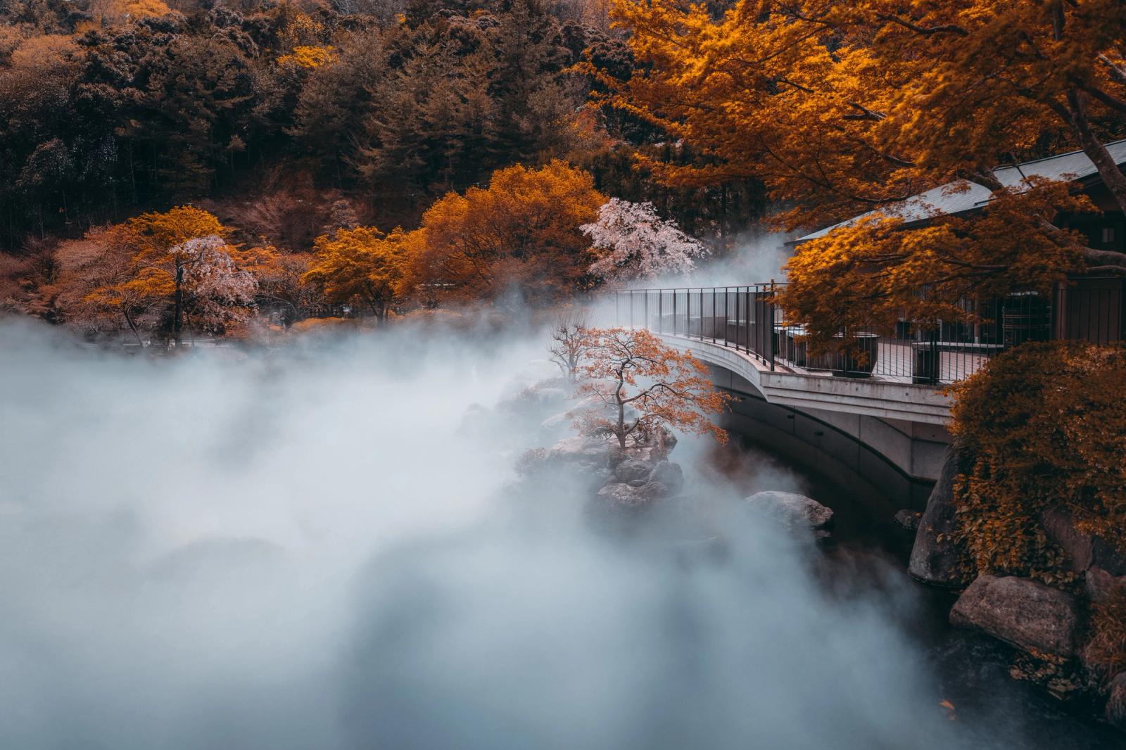 Mist rising over a river beside autumn trees and a covered footbridge