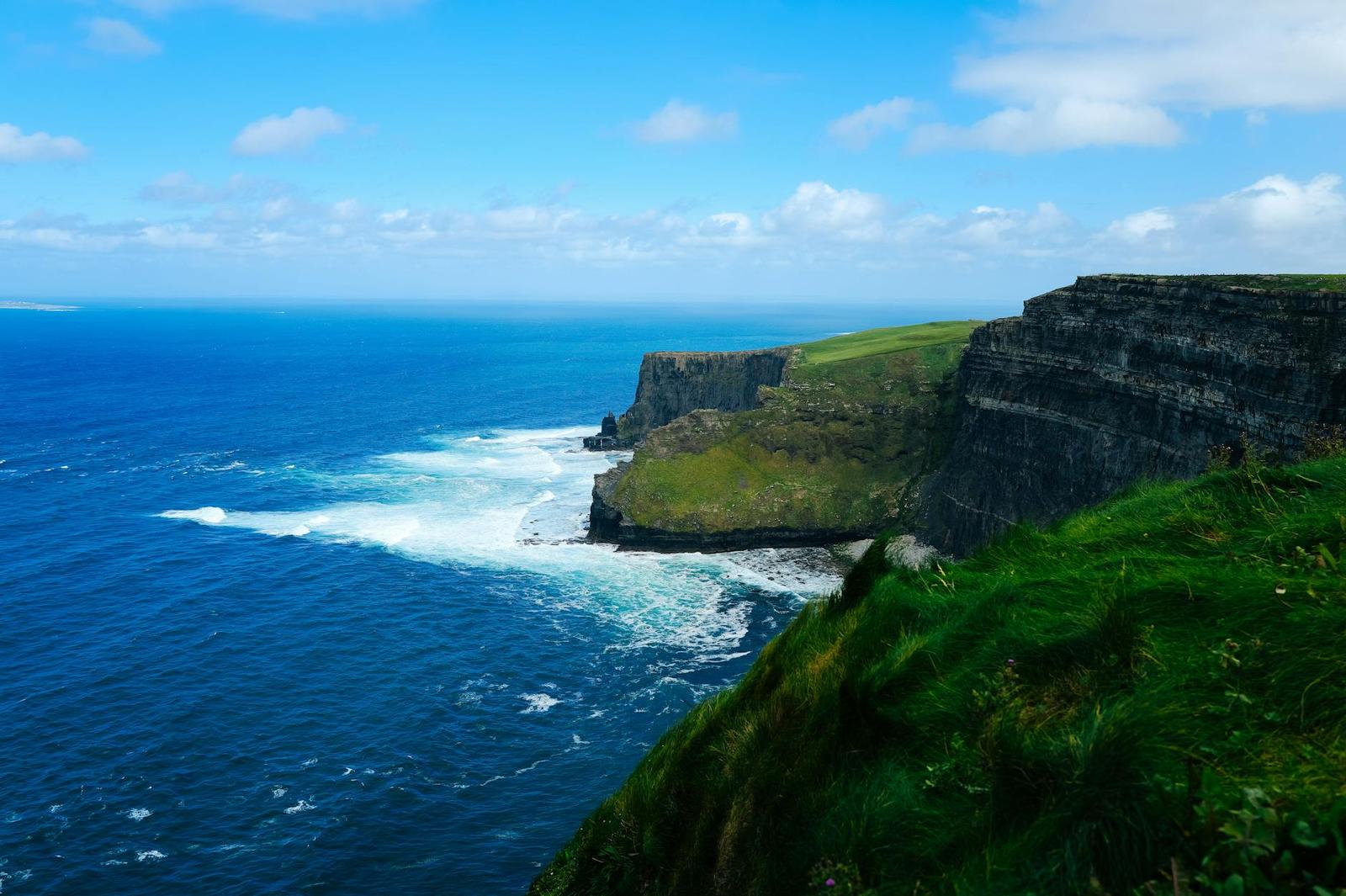 Steep green cliffs plunge into a deep blue ocean under a partly cloudy sky