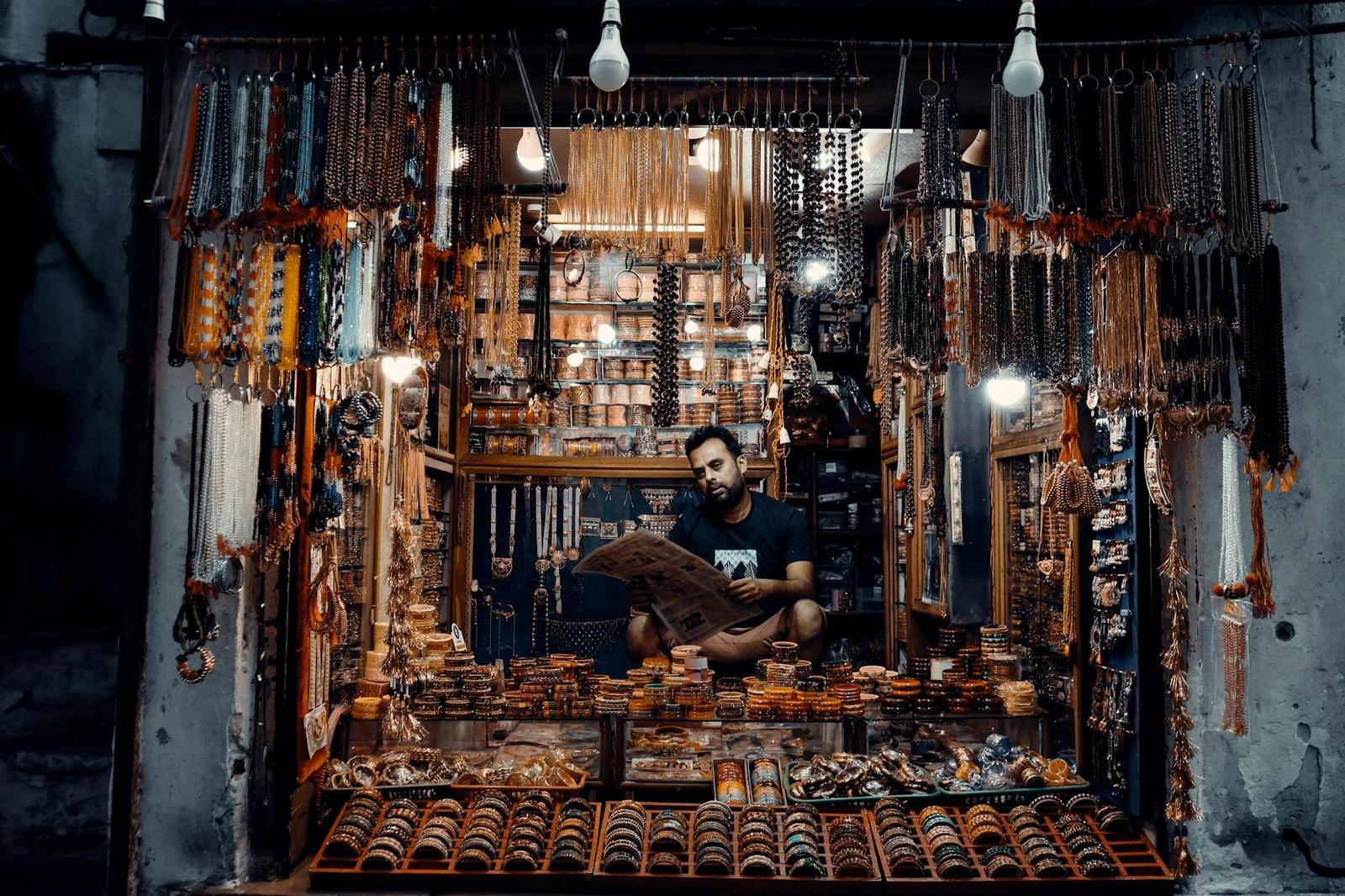 A vendor surrounded by hanging bead necklaces and bracelets in a lit stall