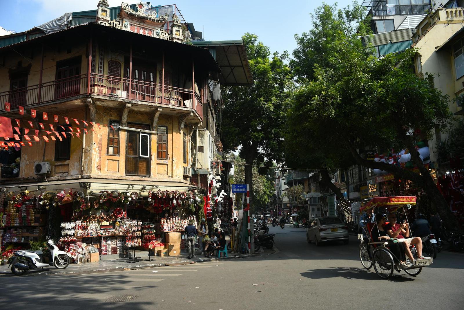 A corner shophouse with a balcony above street stalls and a cyclo parked nearby