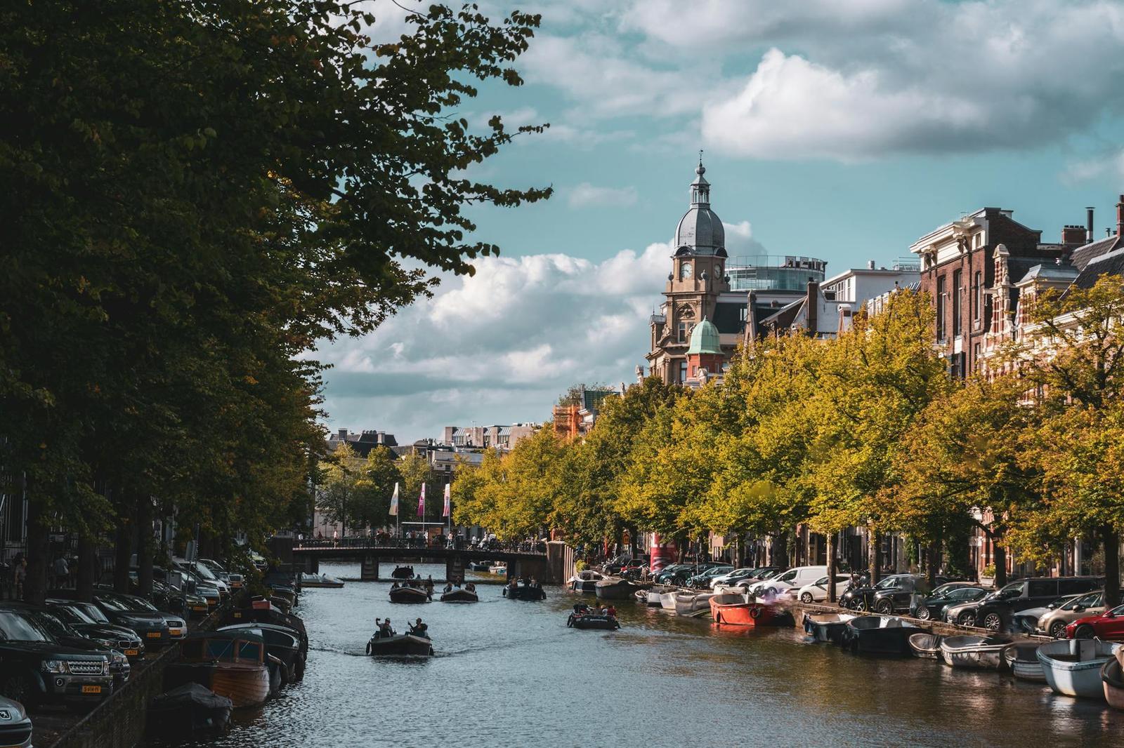 Boats gliding along a tree-lined canal past parked cars and a domed tower