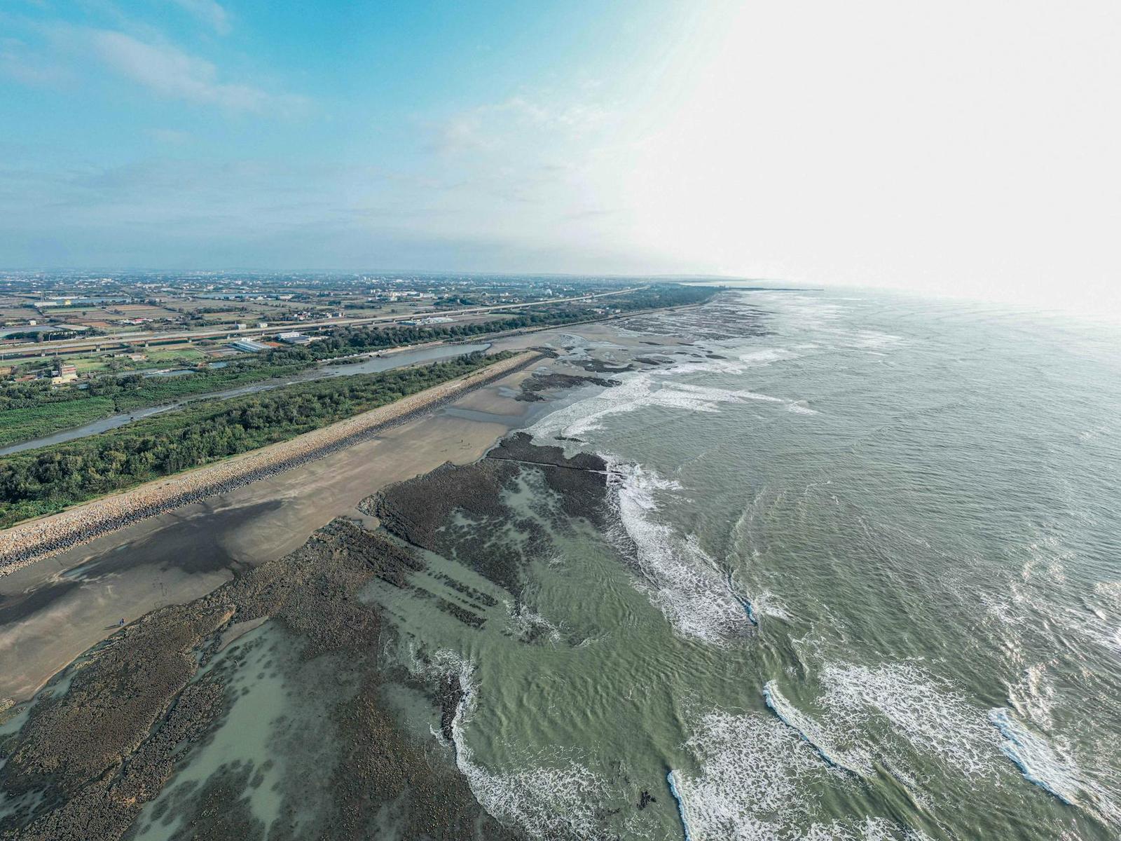 Aerial view of waves breaking along a muddy tidal coastline beside farmland