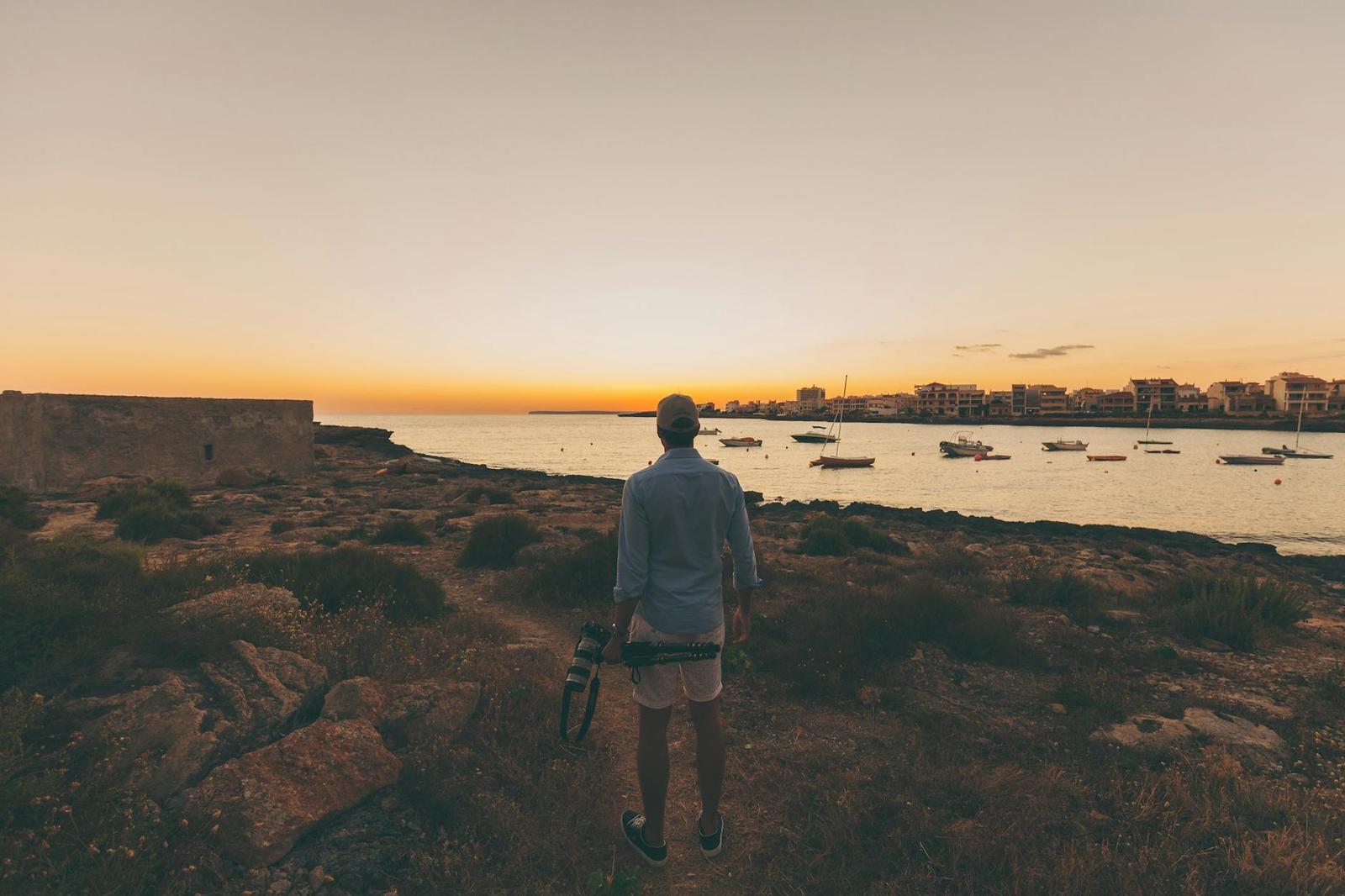 A traveller with a camera looks out over a marina at sunset from a rocky point