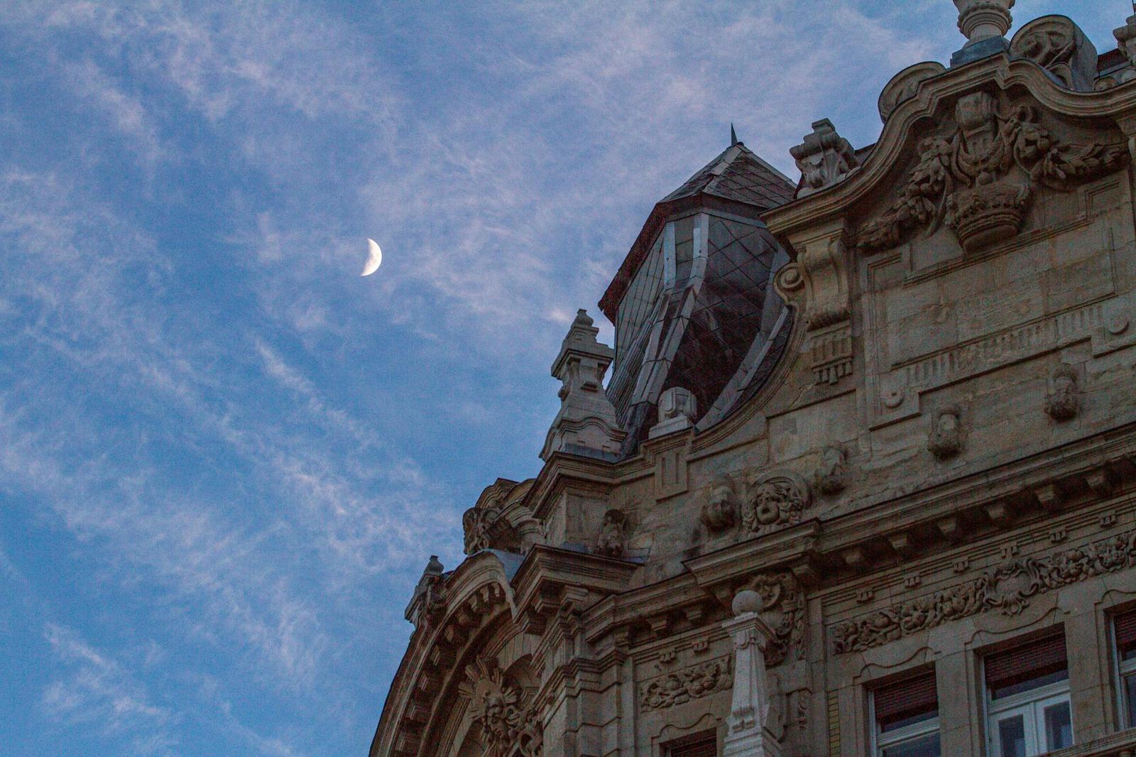 A crescent moon rising beside an ornate stone building facade at dusk