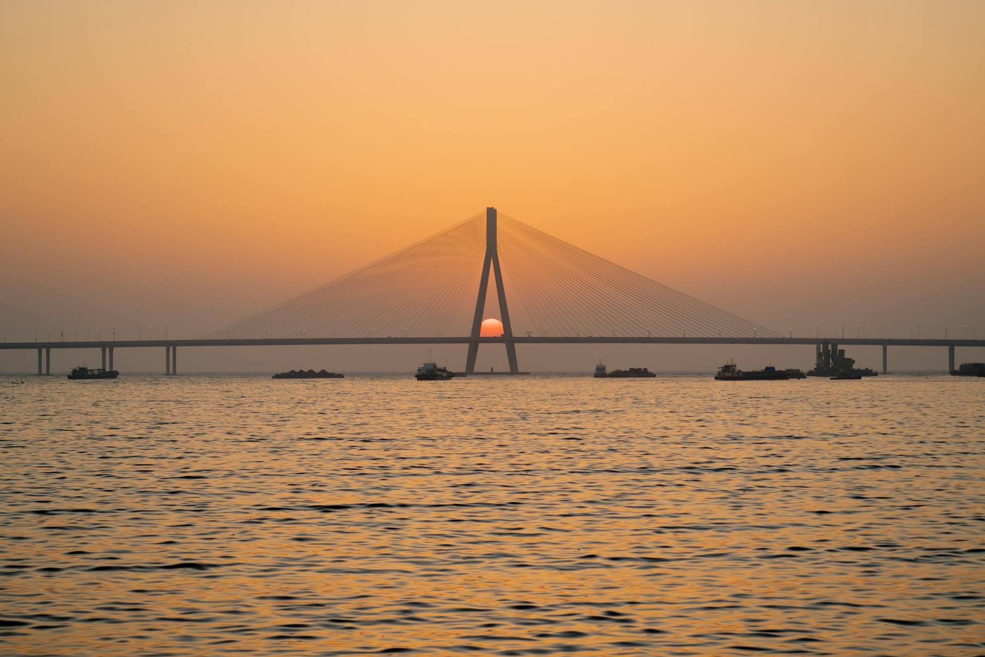 A cable-stayed bridge silhouetted against an orange sunset over water