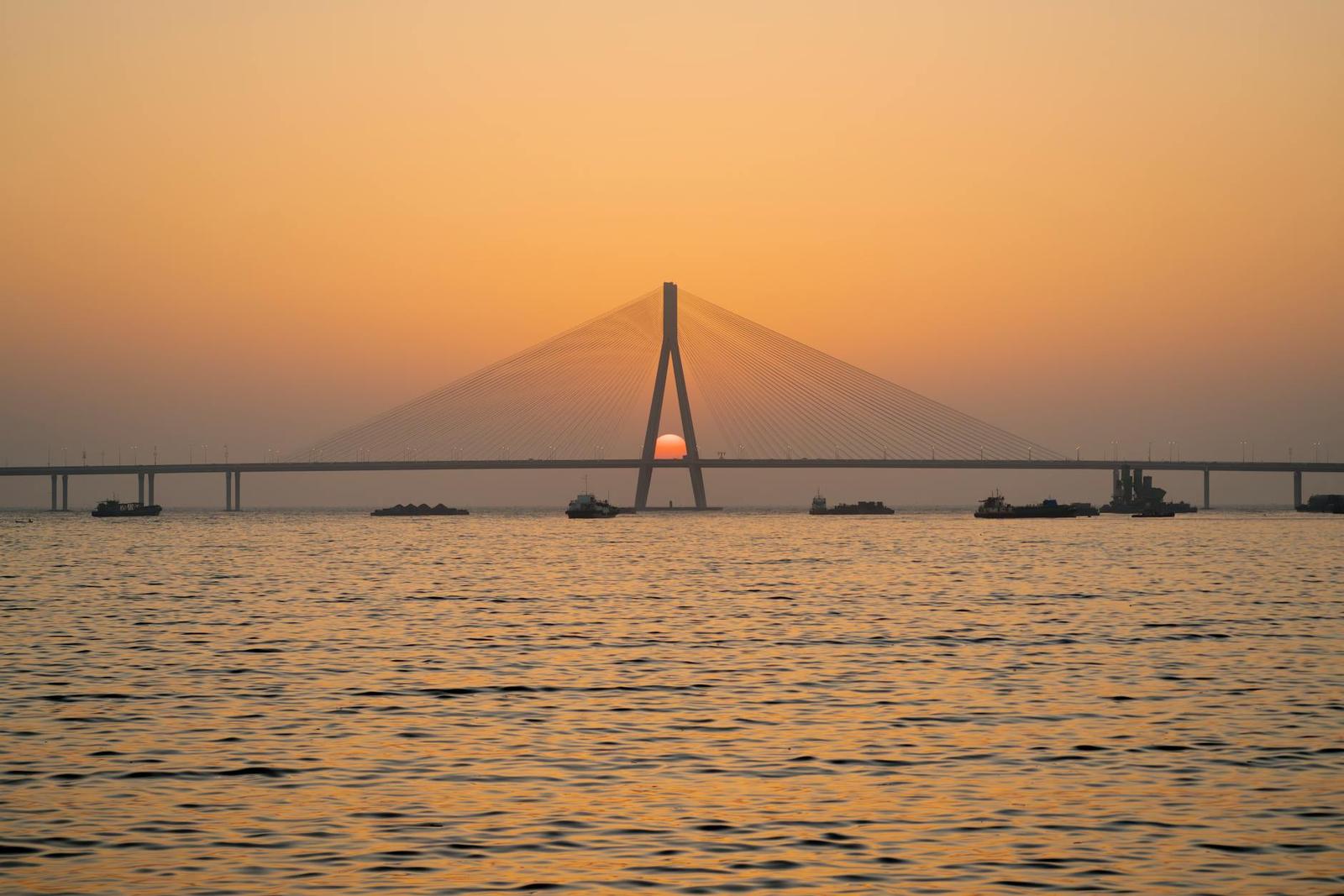 A cable-stayed bridge silhouetted against an orange sunset over water