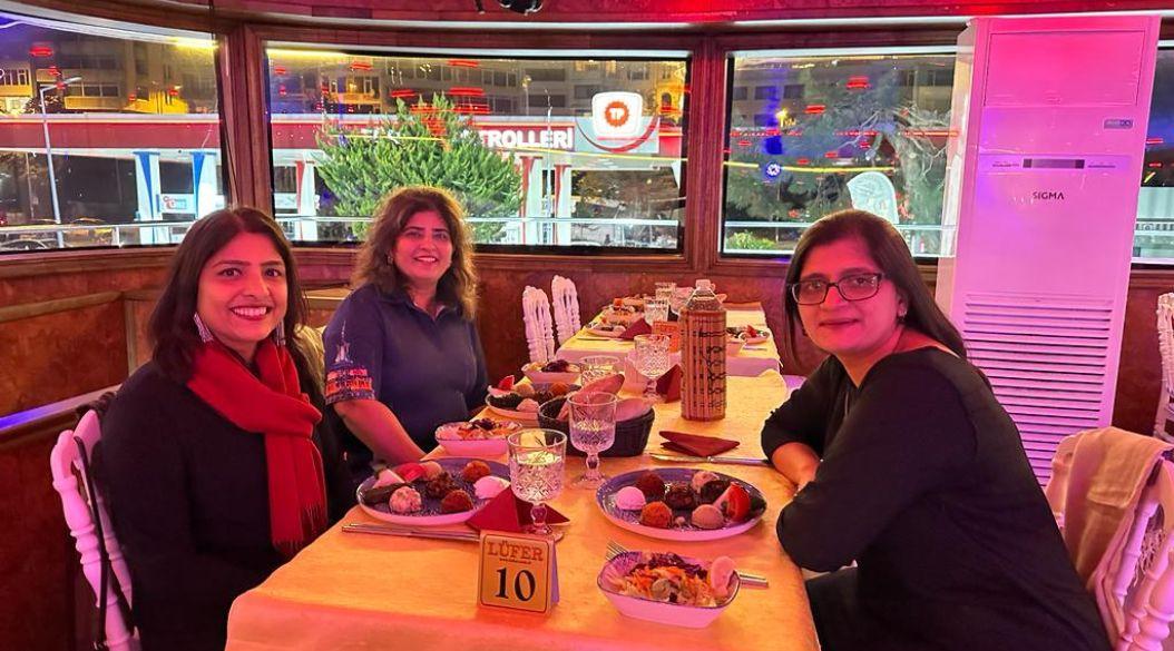 Three women enjoying dinner on a cruise with city lights outside.