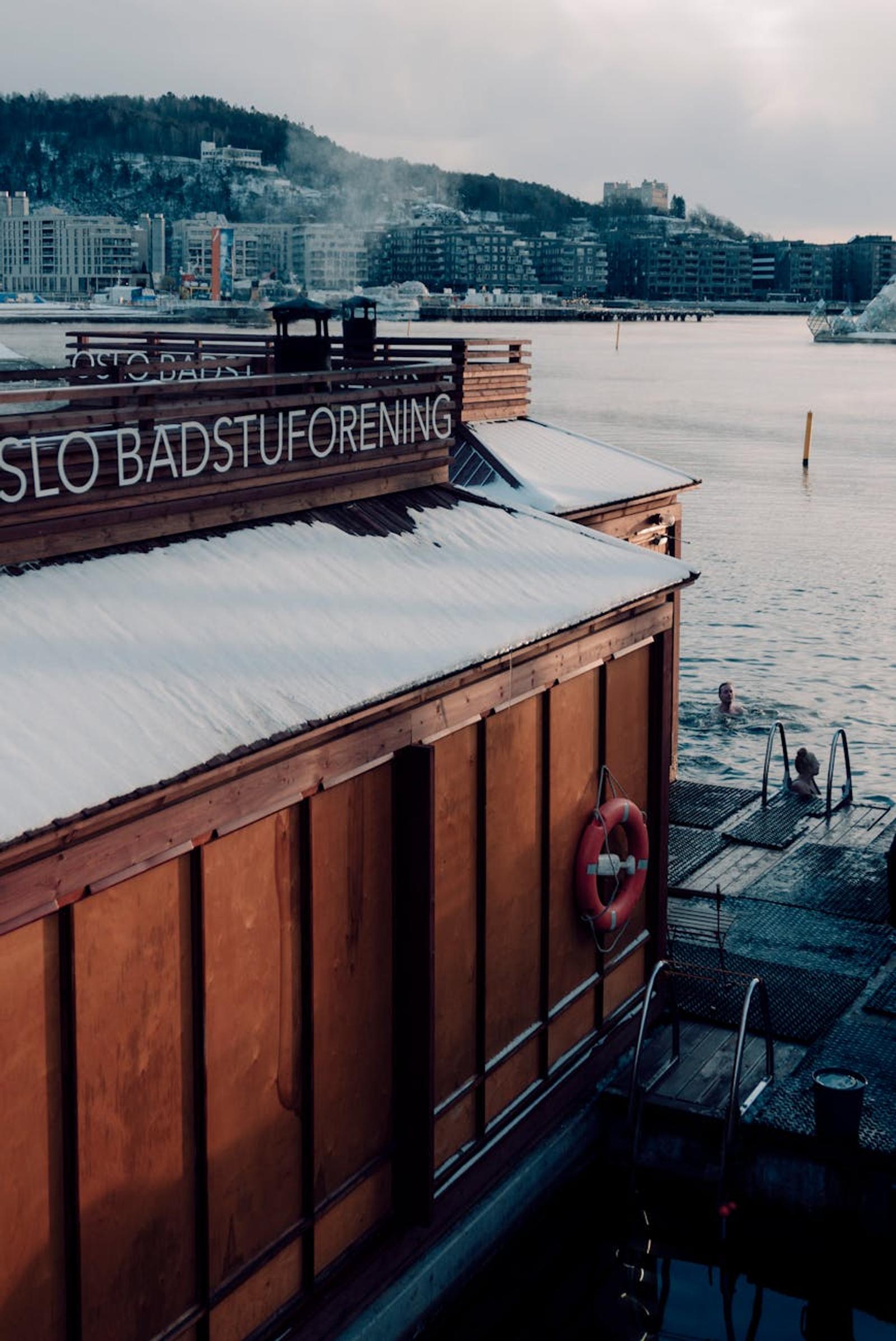 Snow-dusted waterfront sauna cabin with swimmers on a jetty