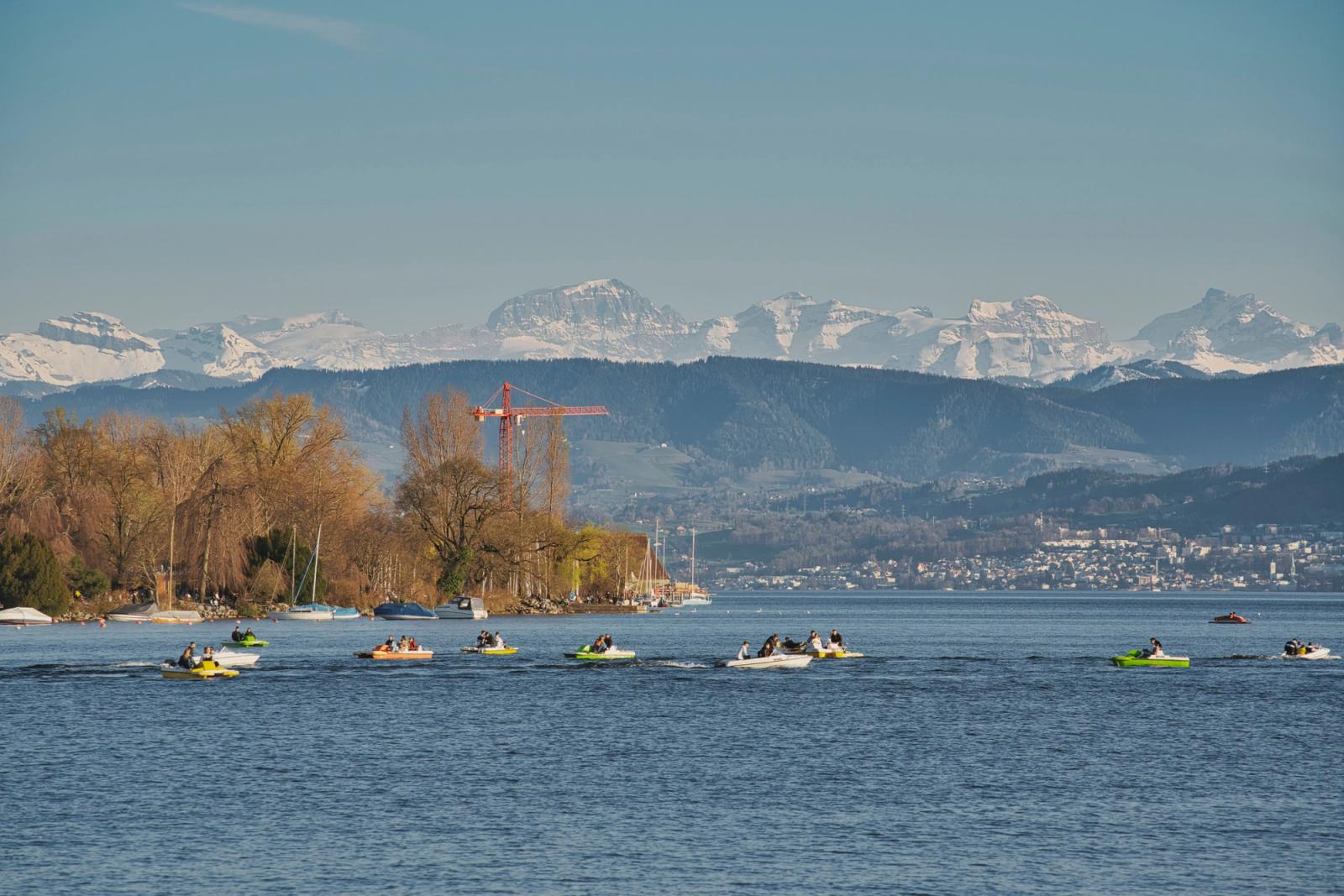 A picturesque view of Lake Zurich with people boating and the snowy Alps in the background