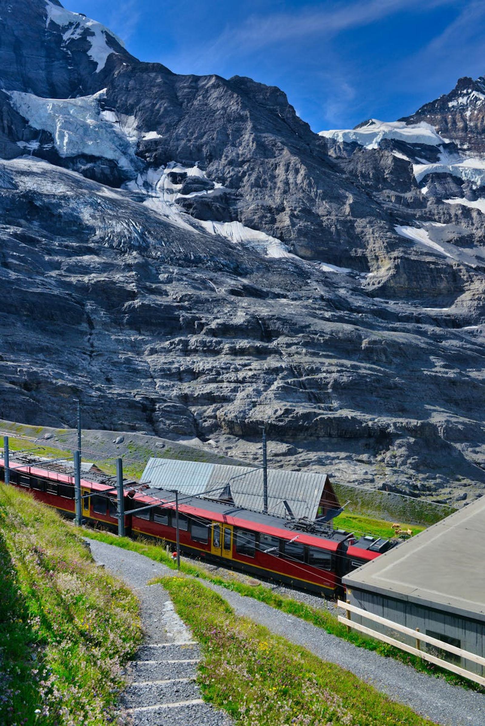 Red cogwheel train descending past a glacier-streaked mountainside