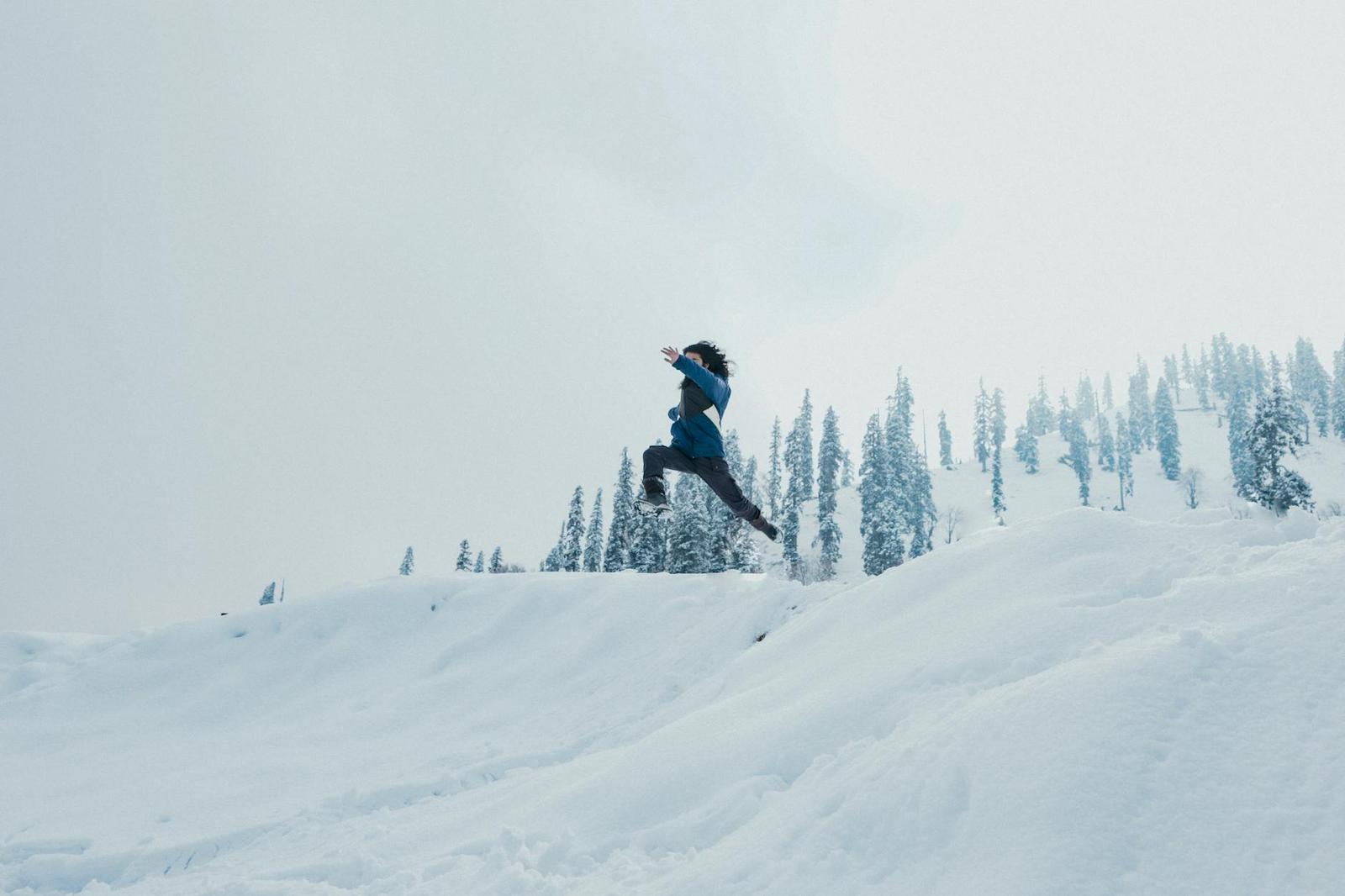 A man leaps across a snow-covered hillside against a backdrop of trees in Gulmarg