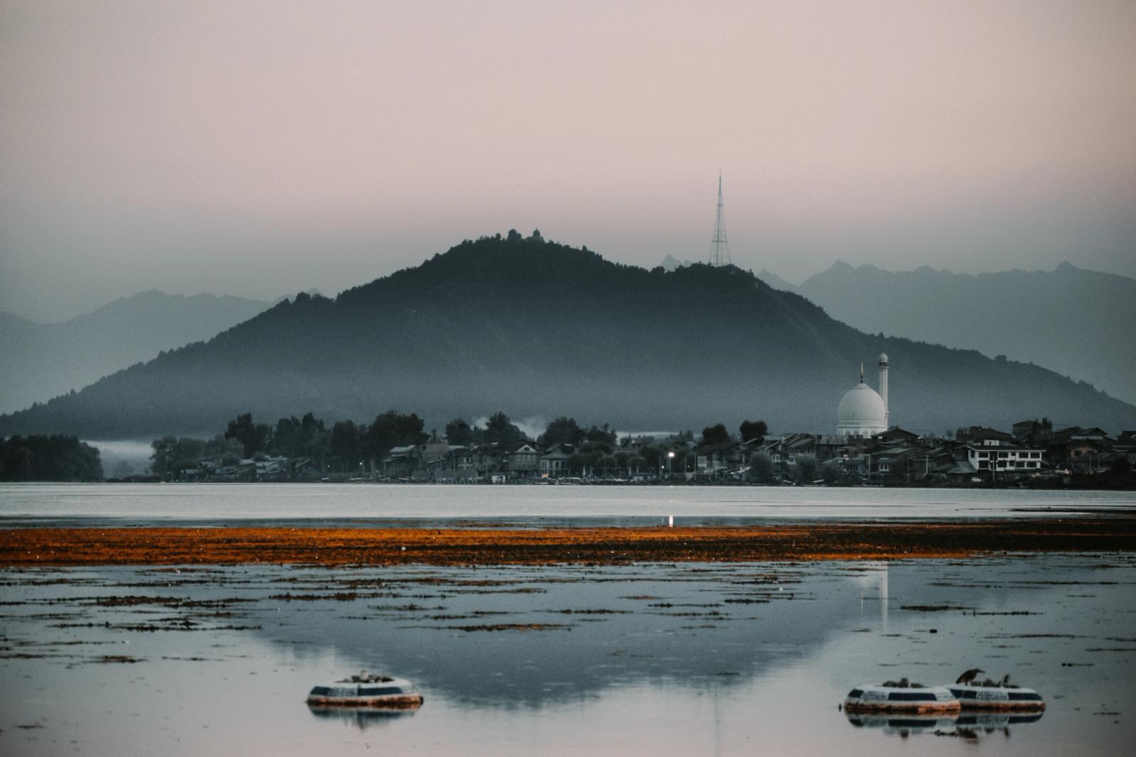Houseboats on a calm lake below a hazy hill and mosque dome at dusk